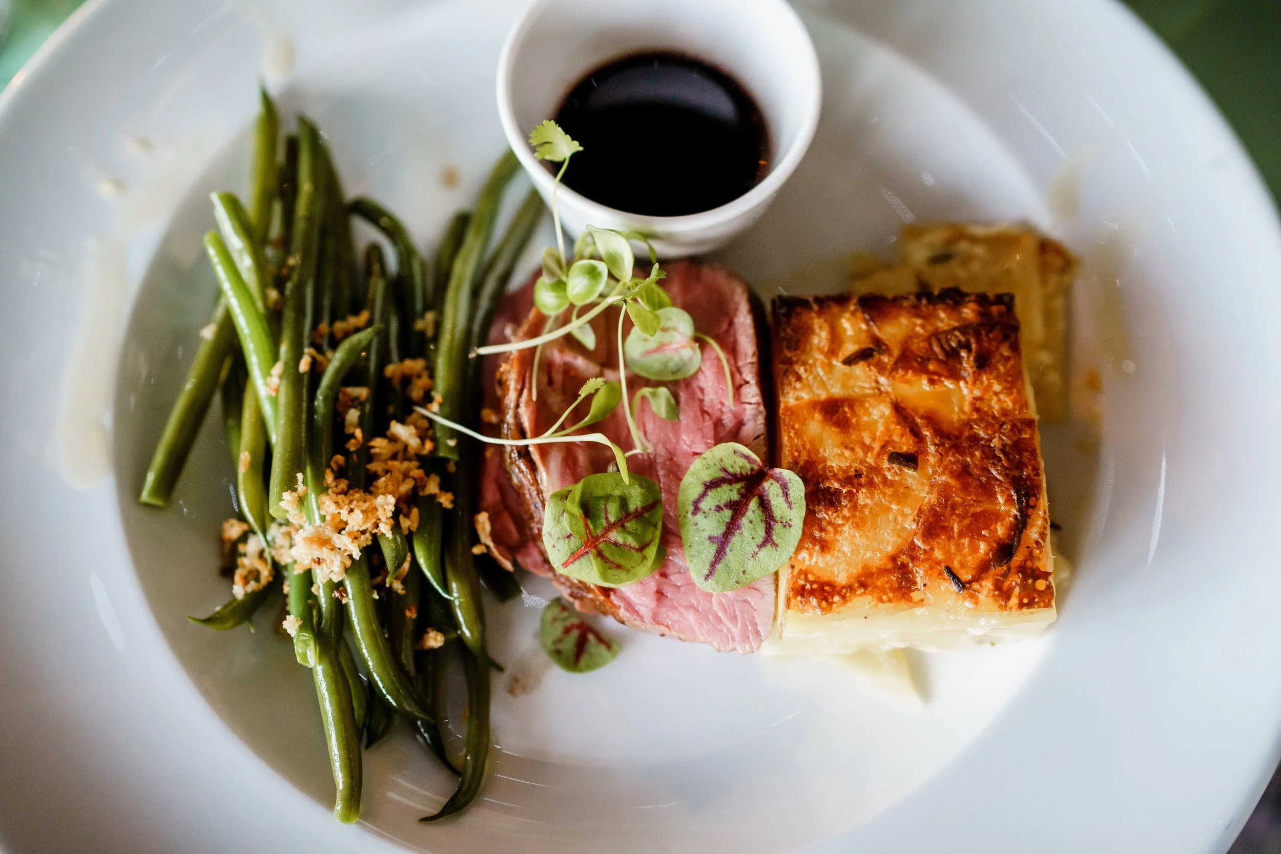 Plate with cooked green beans topped with breadcrumbs, a slice of pink meat, microgreens, a piece of baked salmon with a crispy crust, and a small cup of dark sauce.