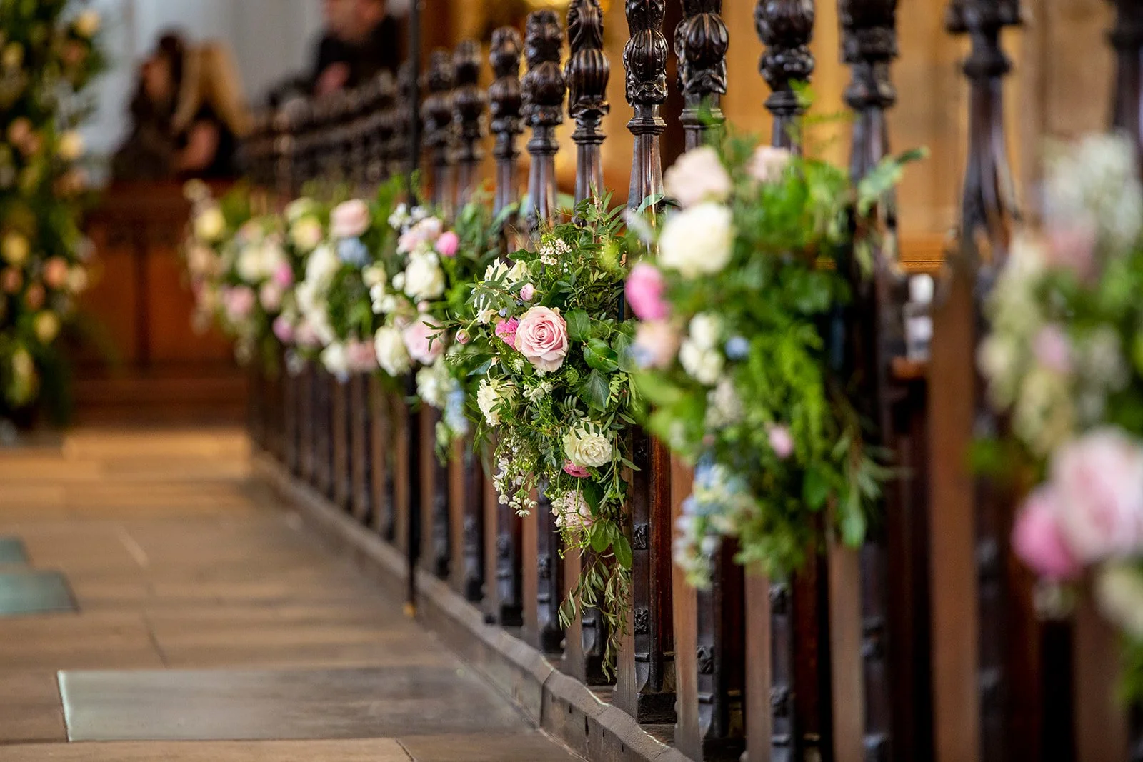 A festive indoor setting with black wrought iron railing and floral arrangements of pink, white, and light blue roses, along with green foliage, decorating the railing, with people in the background.