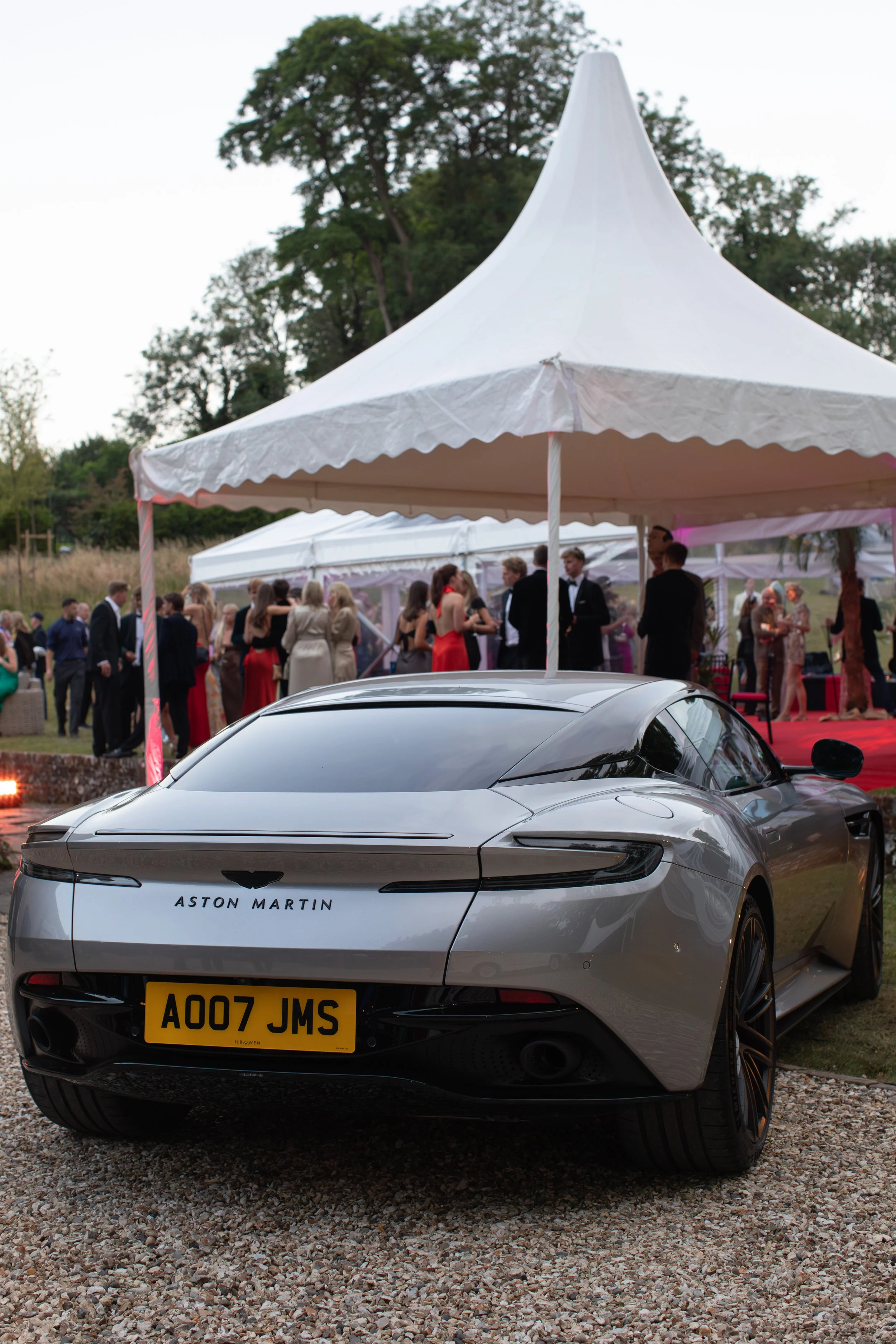 A silver Aston Martin sports car parked on gravel in the foreground with a large white event tent and people dressed formally in the background at an outdoor event.