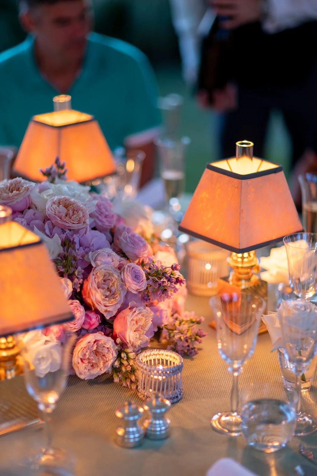 Elegant dining table decorated with pink and purple flowers, warm table lamps, candle holder, and glassware.