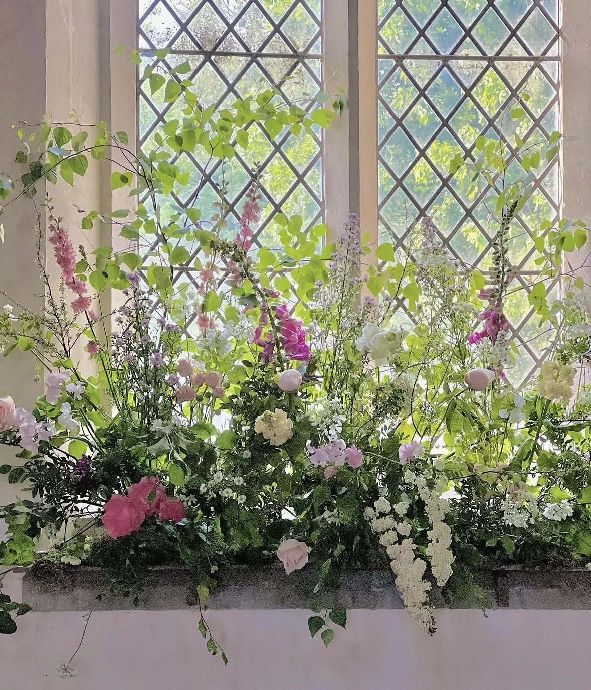 A colorful flower arrangement in a window box with green leafy plants and pink, white, and purple flowers in front of a lattice window.