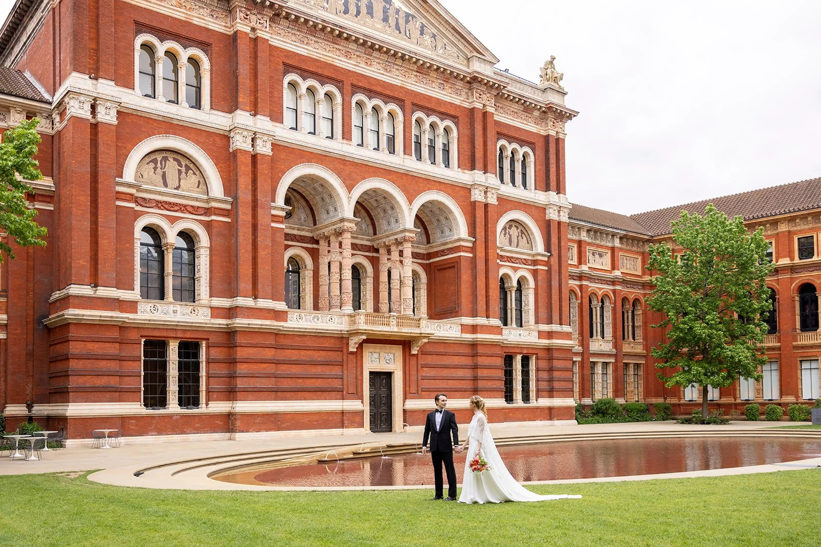 Bride and groom in wedding attire holding hands outside a large red brick building with ornate architectural details, a fountain, and trees.