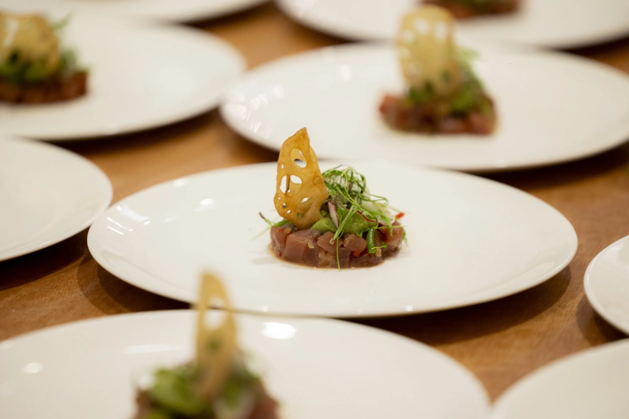 A plated appetizer featuring raw fish, microgreens, a fried lotus root chip, and a dollop of green puree, with additional plates of similar dishes blurred in the background.