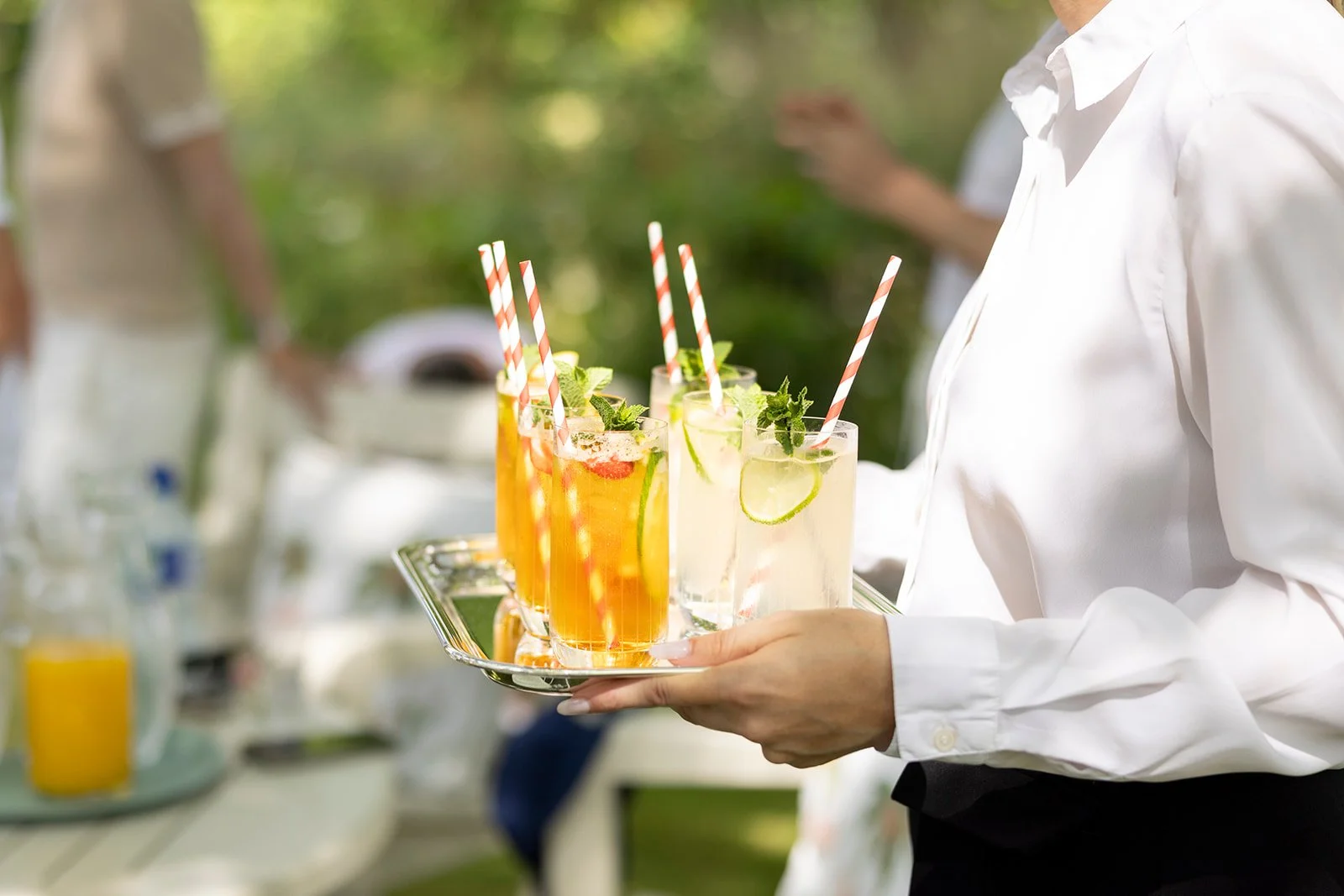 A person holding a tray with several glasses of colorful drinks garnished with mint leaves and slices of lime, at an outdoor event.
