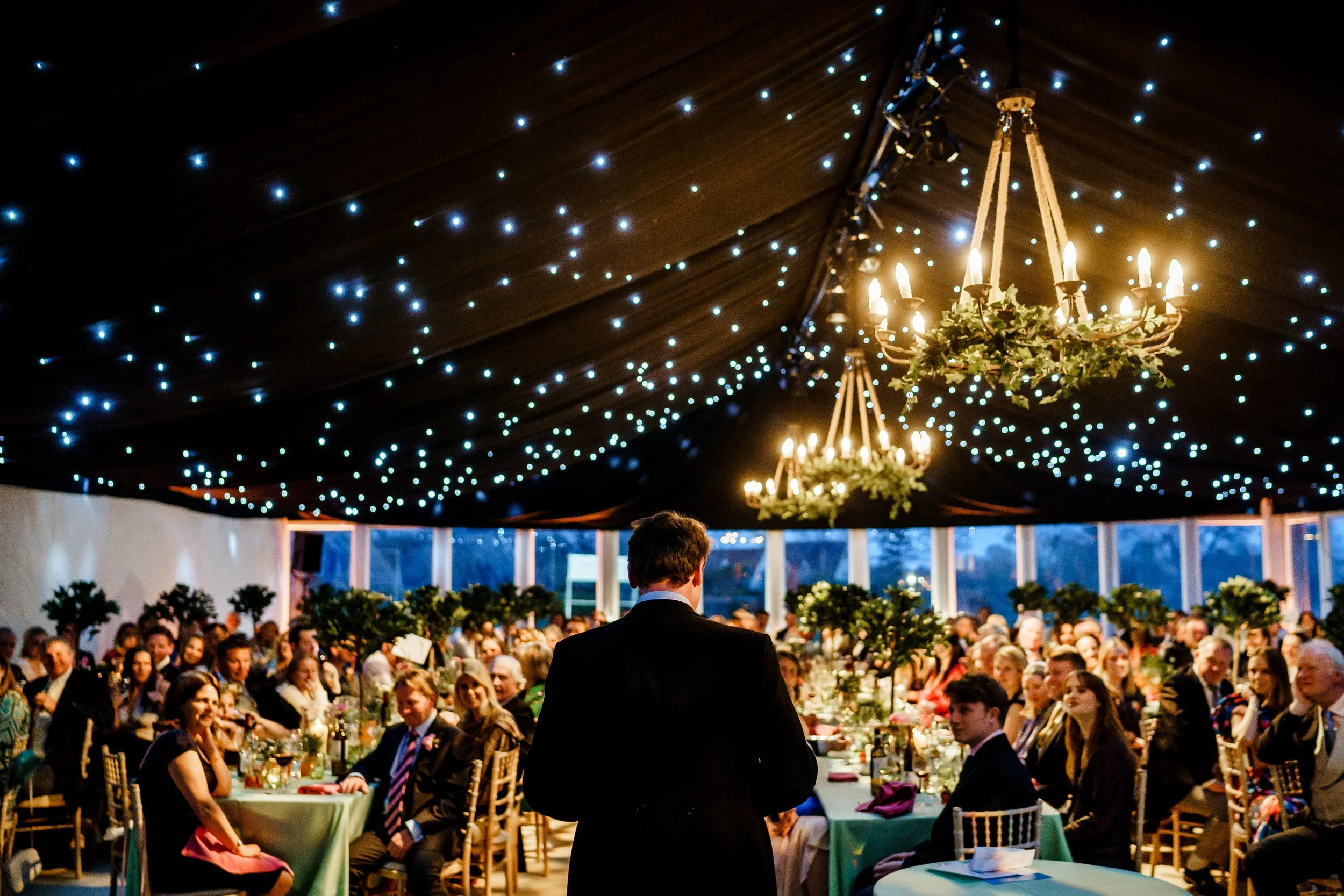 People attending a wedding reception in a decorated tent with string lights and chandeliers, with a man in formal attire standing at the center facing the crowd.
