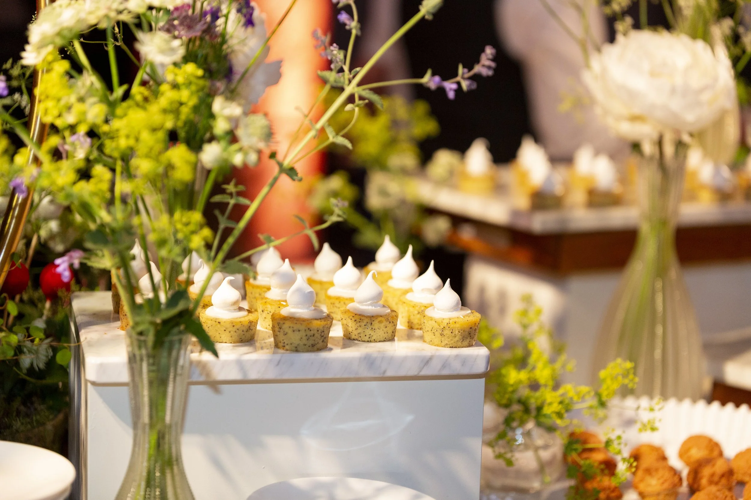 Mini yellow cakes topped with white frosting dots, displayed on a multi-tiered white stand with floral arrangements in the foreground.