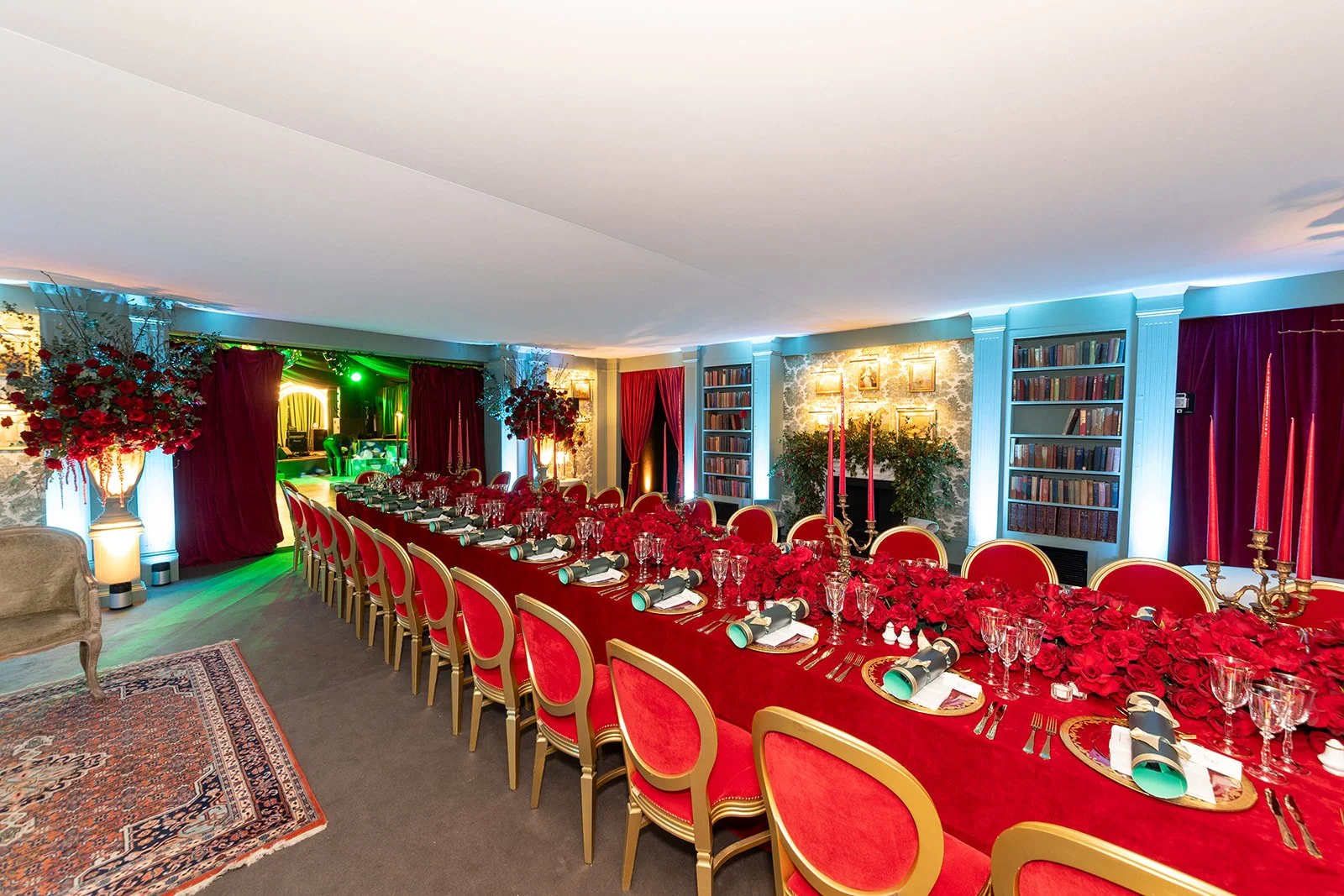 Elegant banquet table set with red roses, candles, glassware, and silverware in a decorated dining room with bookshelves and curtains.