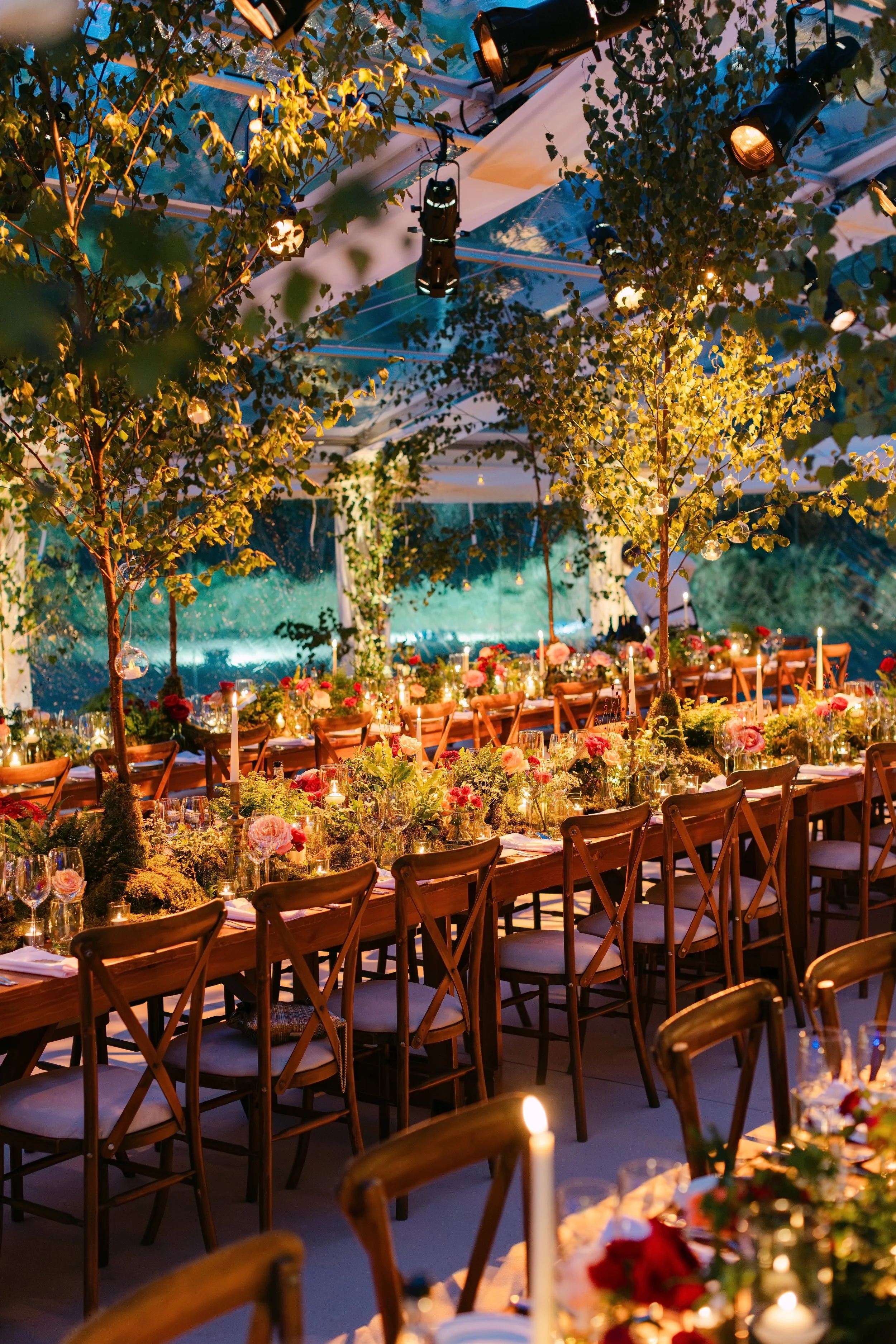 A festive indoor dining setup with wooden chairs and a long table decorated with flowers, candles, and greenery. The ceiling is adorned with hanging trees, string lights, and professional stage lighting, creating a warm, whimsical atmosphere.