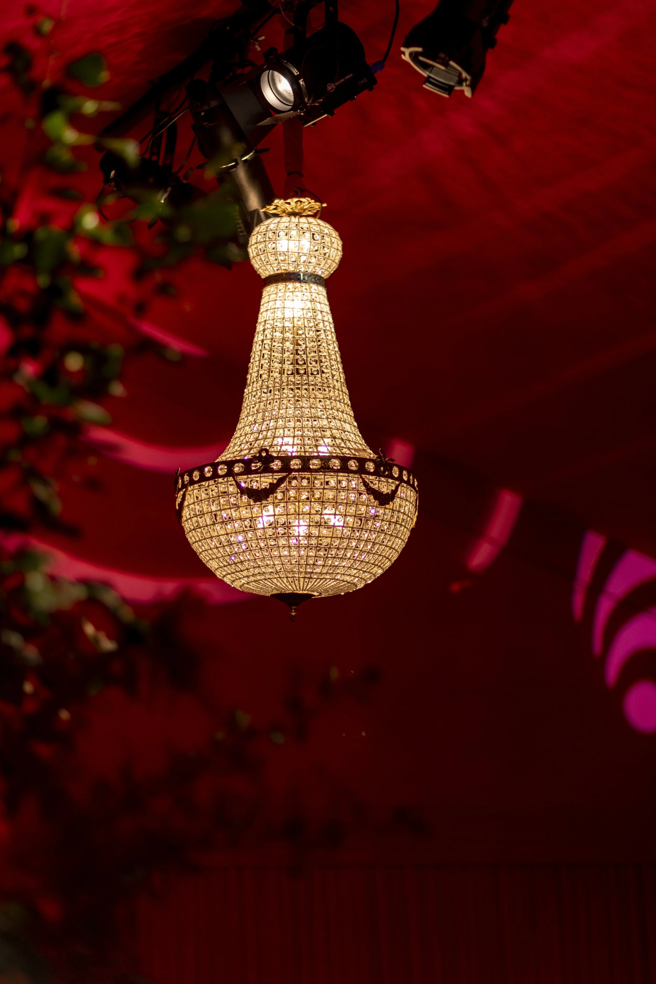 A decorative chandelier hanging from a ceiling with red lighting, with some greenery partially visible in the foreground.