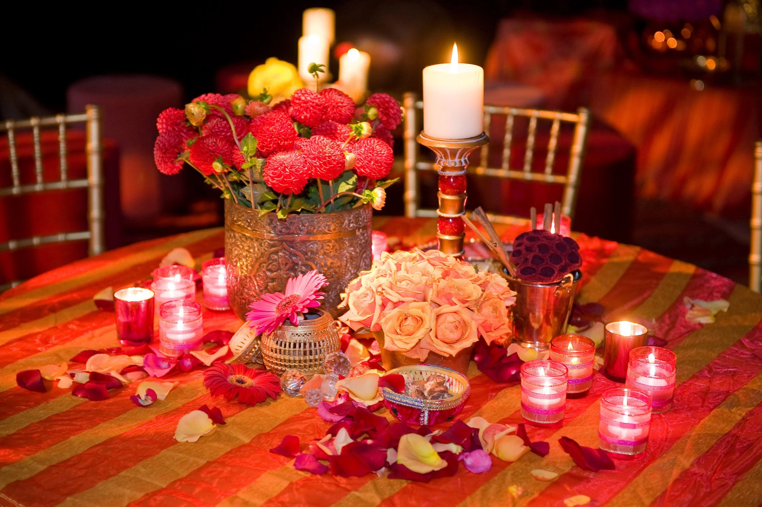 A table decorated with red and pink flowers, lit candles, and scattered flower petals, likely for a romantic or celebration occasion.
