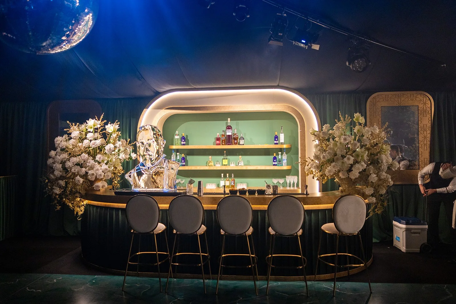 A contemporary bar setup with a rounded counter, chairs, and artworks. The bar has a green background with bottles displayed on shelves, surrounded by floral arrangements and a metallic sculpture. There are people on the right side, one adjusting something, and a dark ceiling with ceiling lights and a globe reflection.