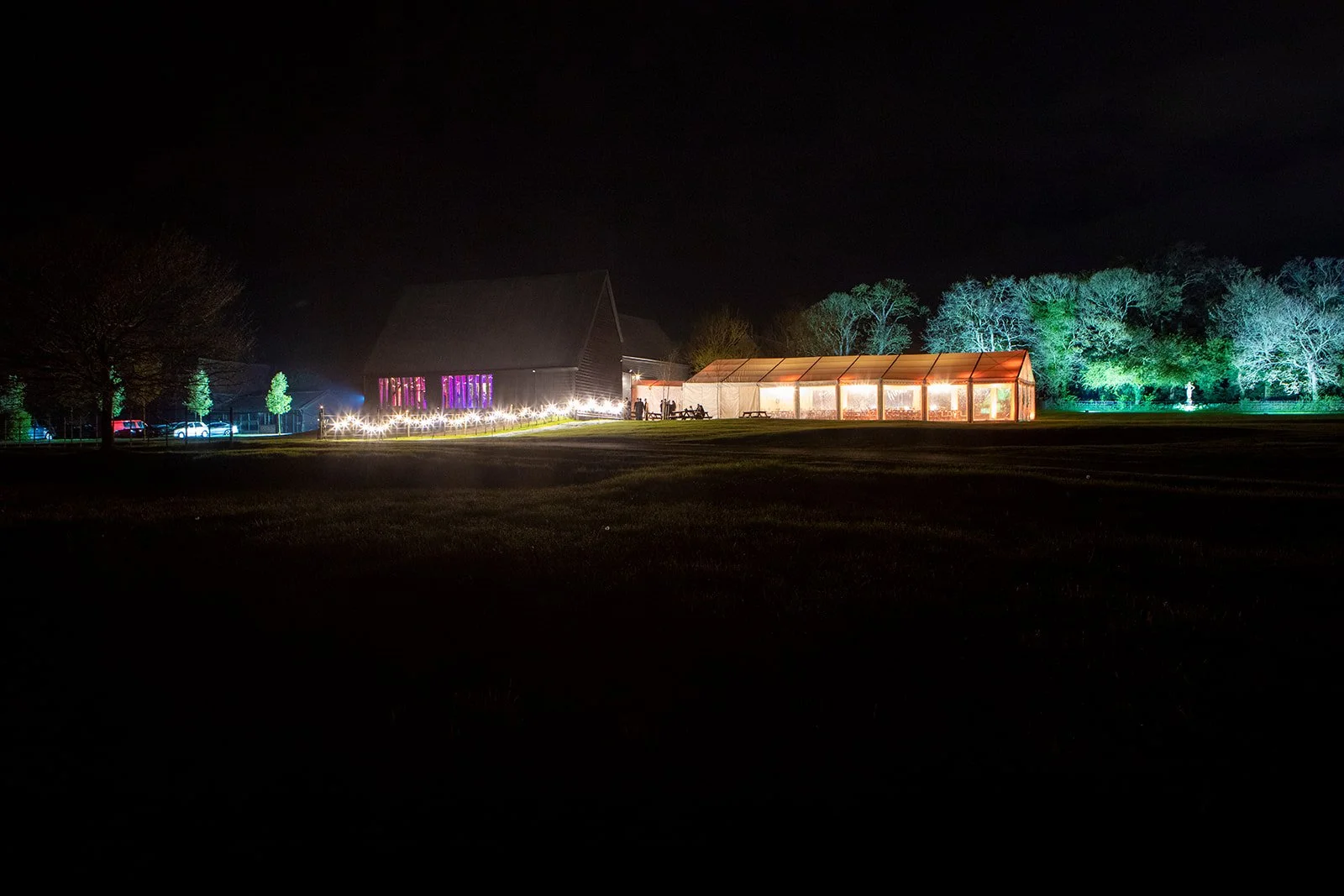 Night view of a lit outdoor event space with a large barn, a tent, trees, and string lights.