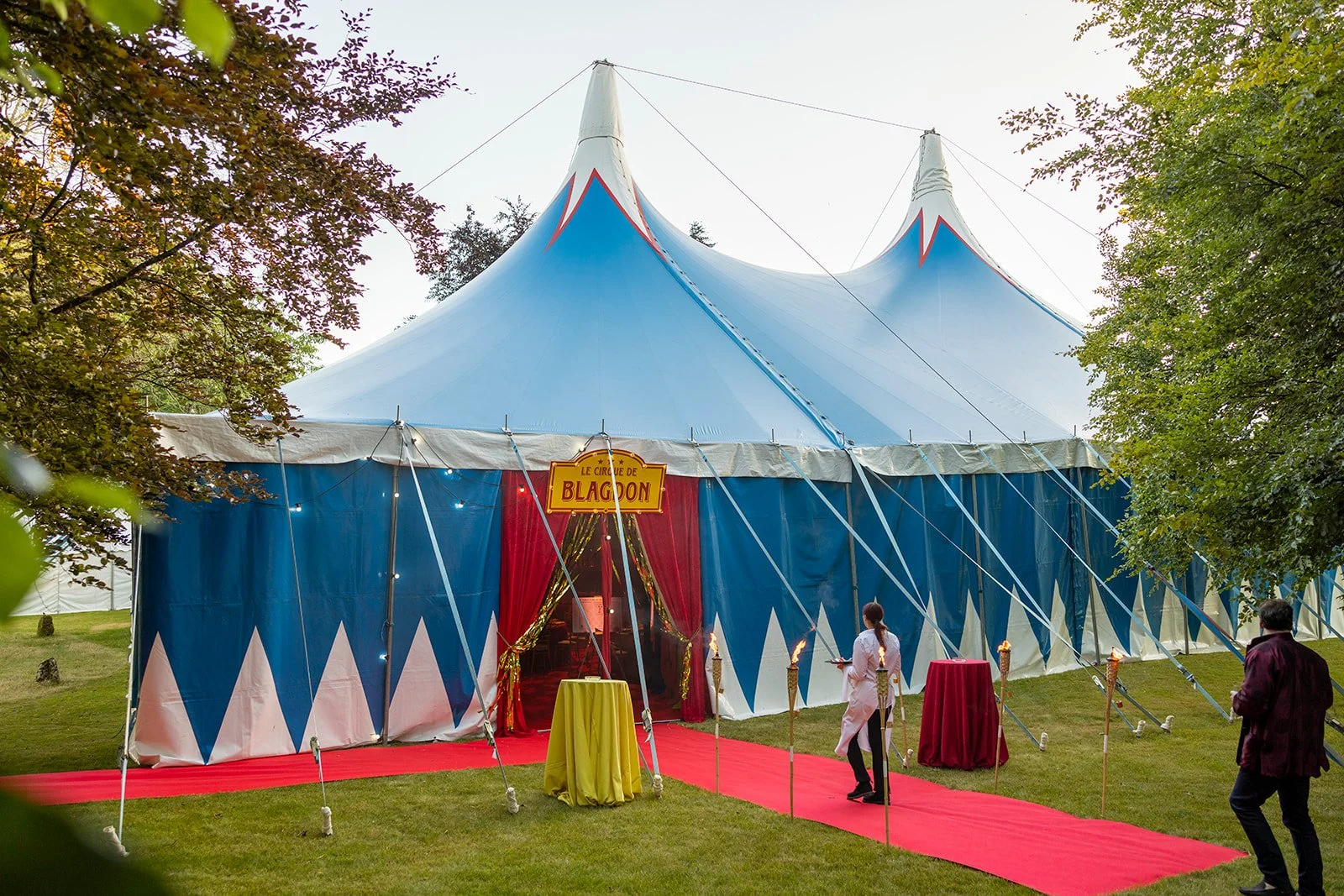 A large blue circus tent with red accents, surrounded by trees, with a red carpet leading to the entrance. There are people near the entrance, and a sign reading 'Le Cirque de Blagoon.'