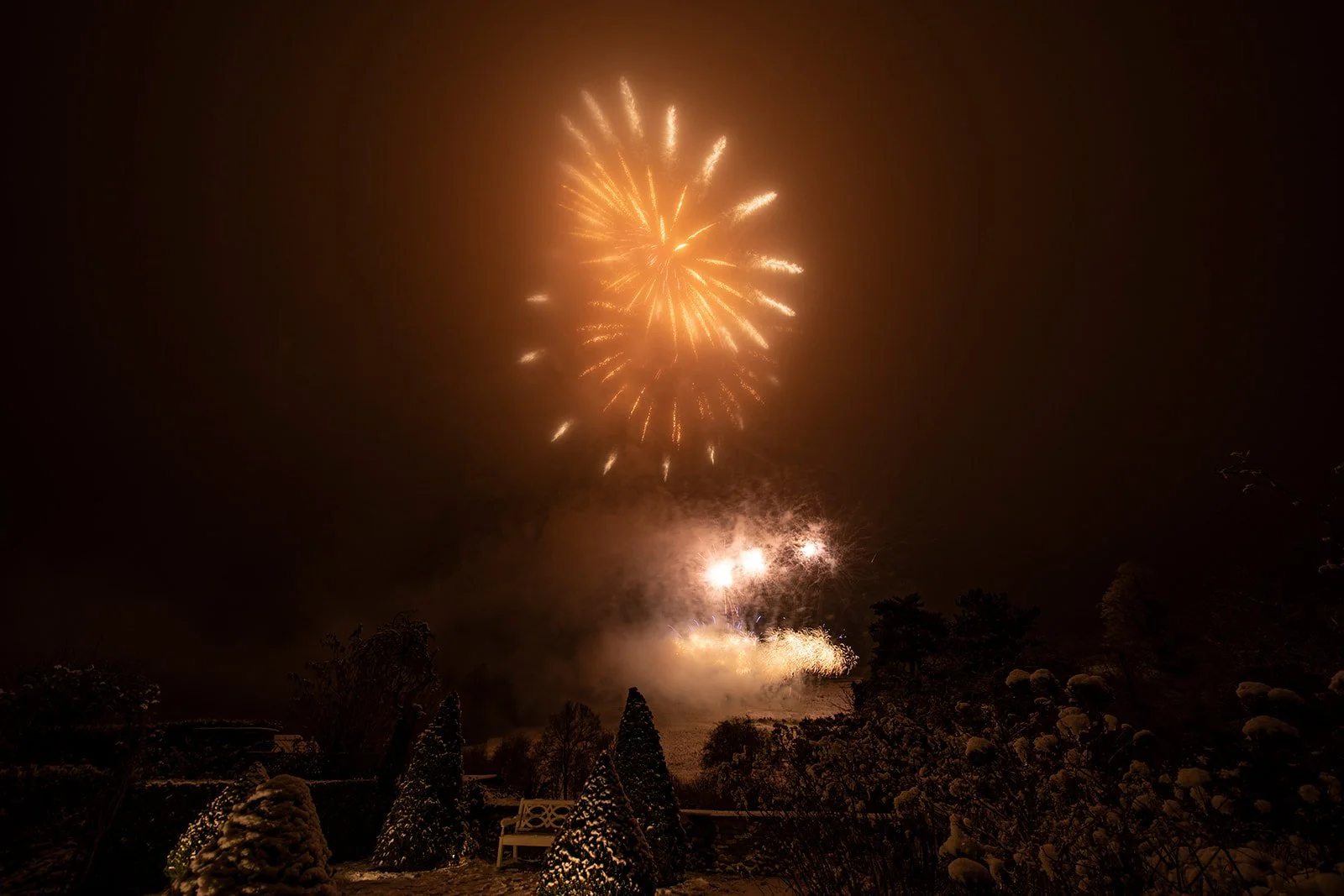 Fireworks light up the night sky over a snowy landscape with trees and a bench in the foreground.