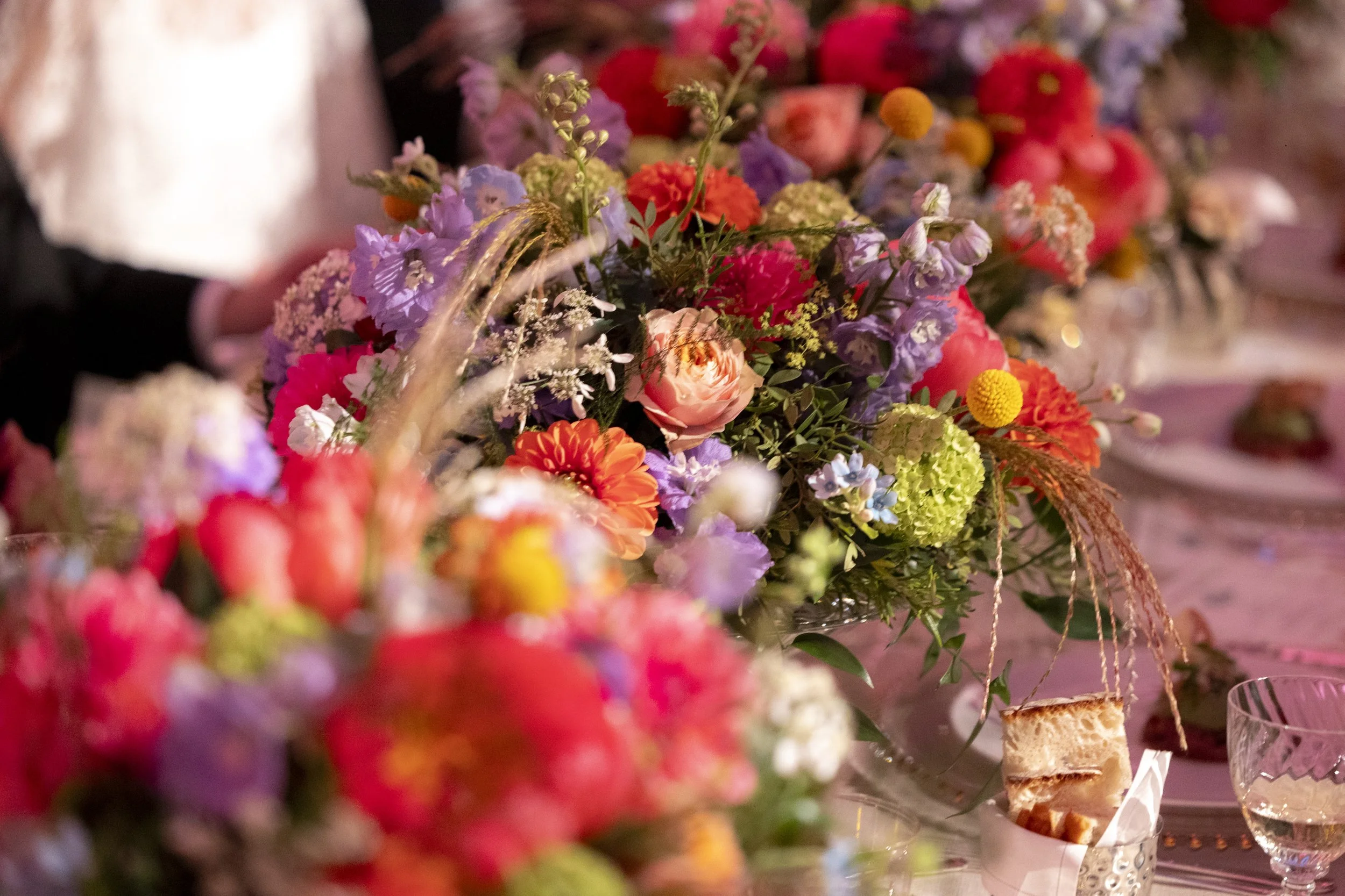 A colorful floral centerpiece on a decorated table featuring a variety of flowers such as pink roses, purple, orange, and yellow blooms, with some food items like bread and cheese visible on the table.