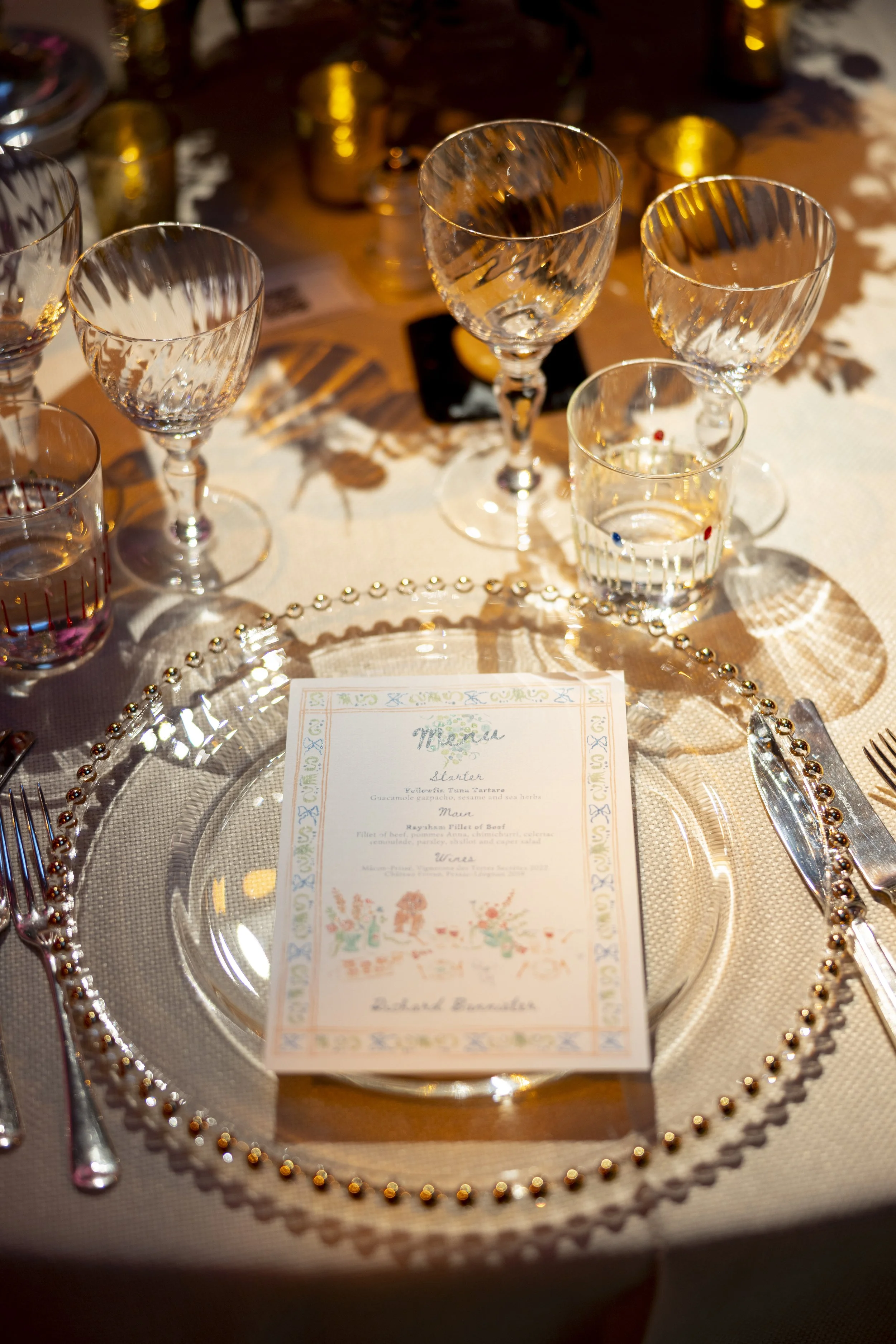 A formal table setting with a glass plate, silverware, and a menu card at the center. Several empty crystal wine glasses are arranged on the table, casting shadows and reflections. The menu card is decorated with colorful illustrations and floral pat