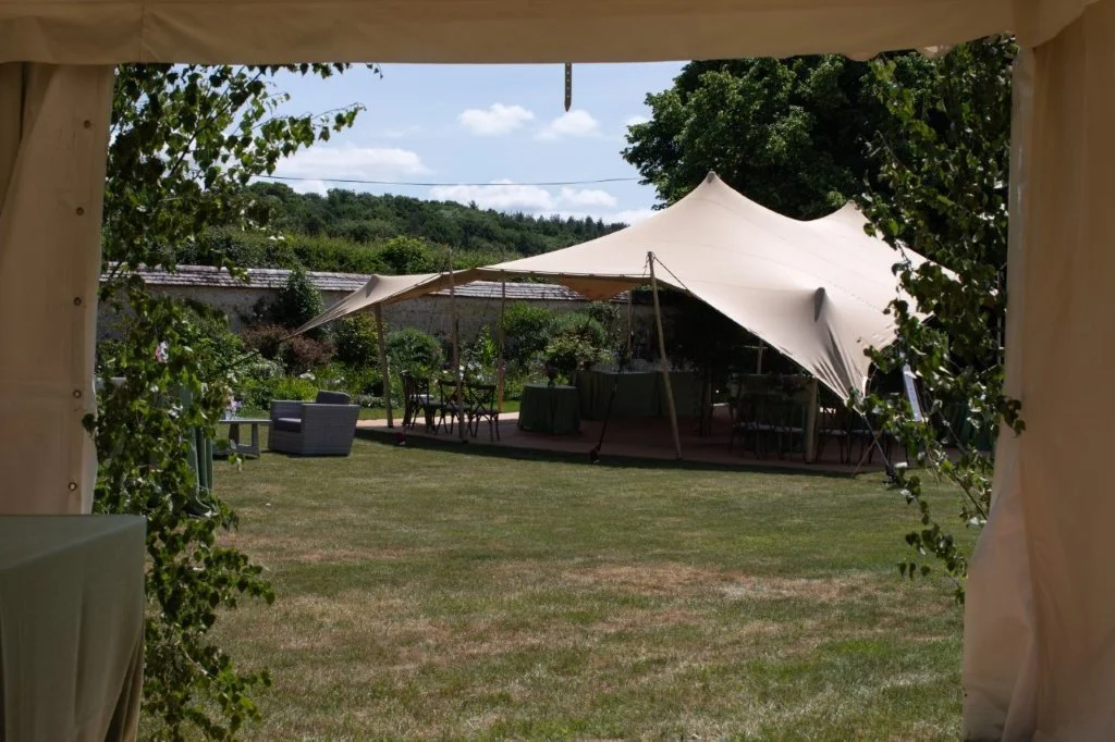Outdoor event setup with a tent, tables, chairs, and lounge furniture in a garden area with trees and a clear sky.