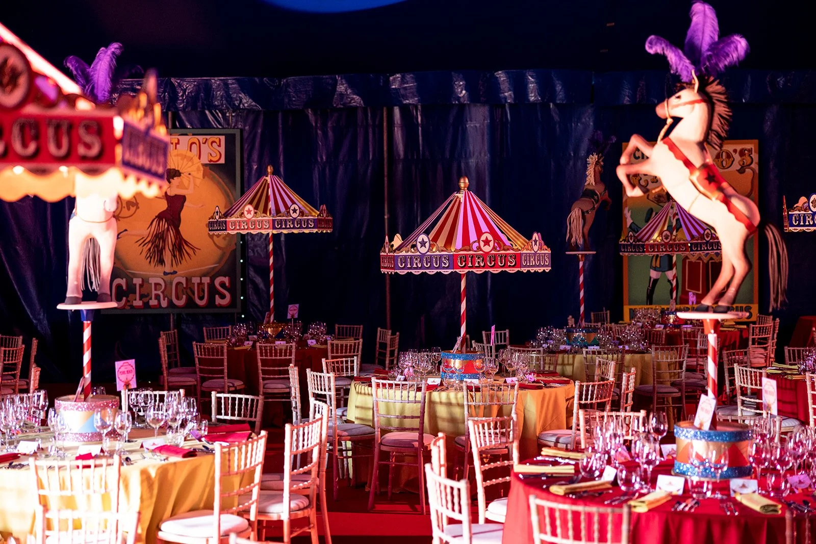 Colorful circus-themed event space with decorated tables, chairs, and vintage circus signs, including carousel horse and clown images, under vibrant striped tents.