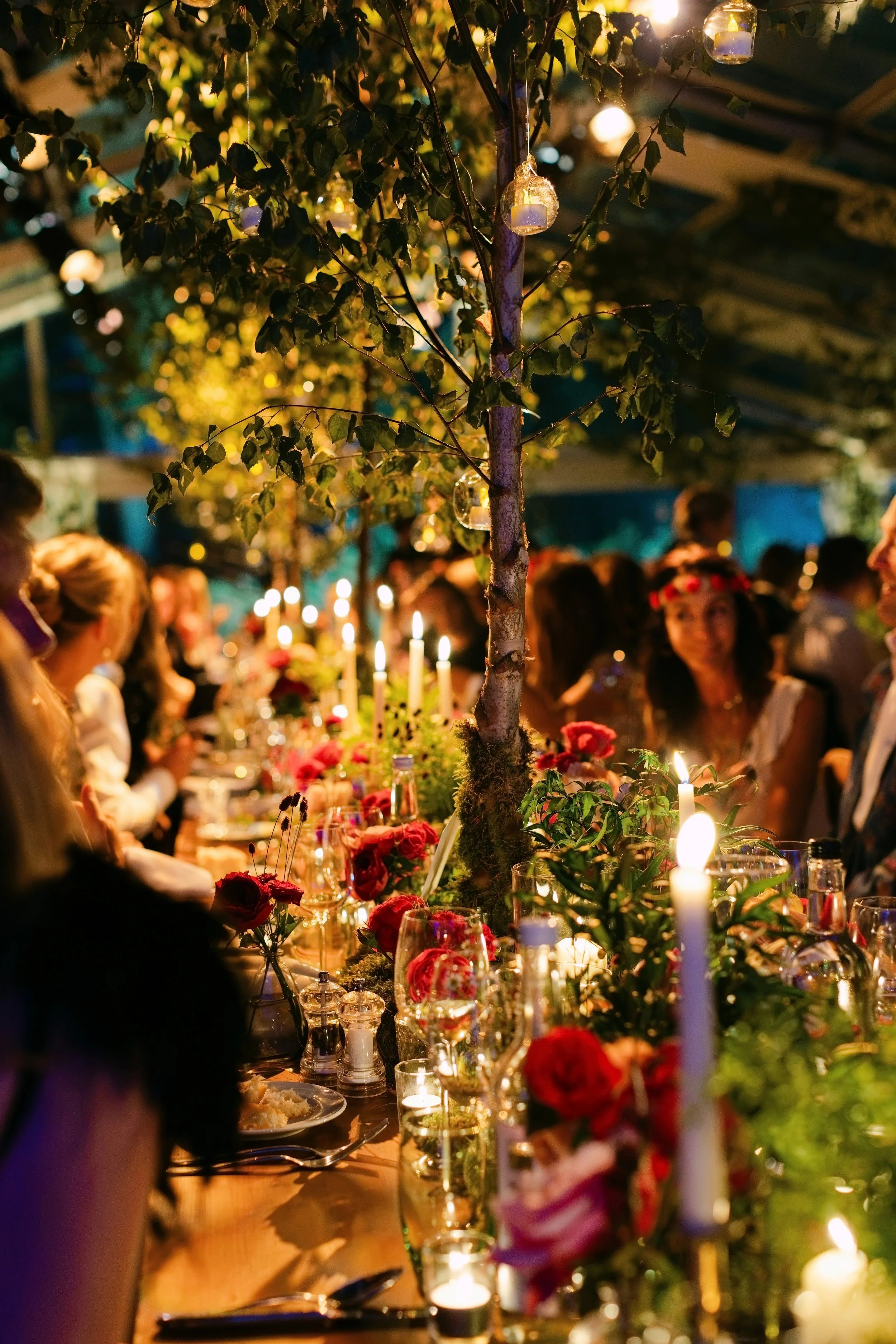 A nighttime indoor party scene with a long dining table decorated with candles, flowers, and greenery. Overhead, fairy lights and hanging jars with candles illuminate the space. Guests are gathered around the table, engaged in conversations and enjoy