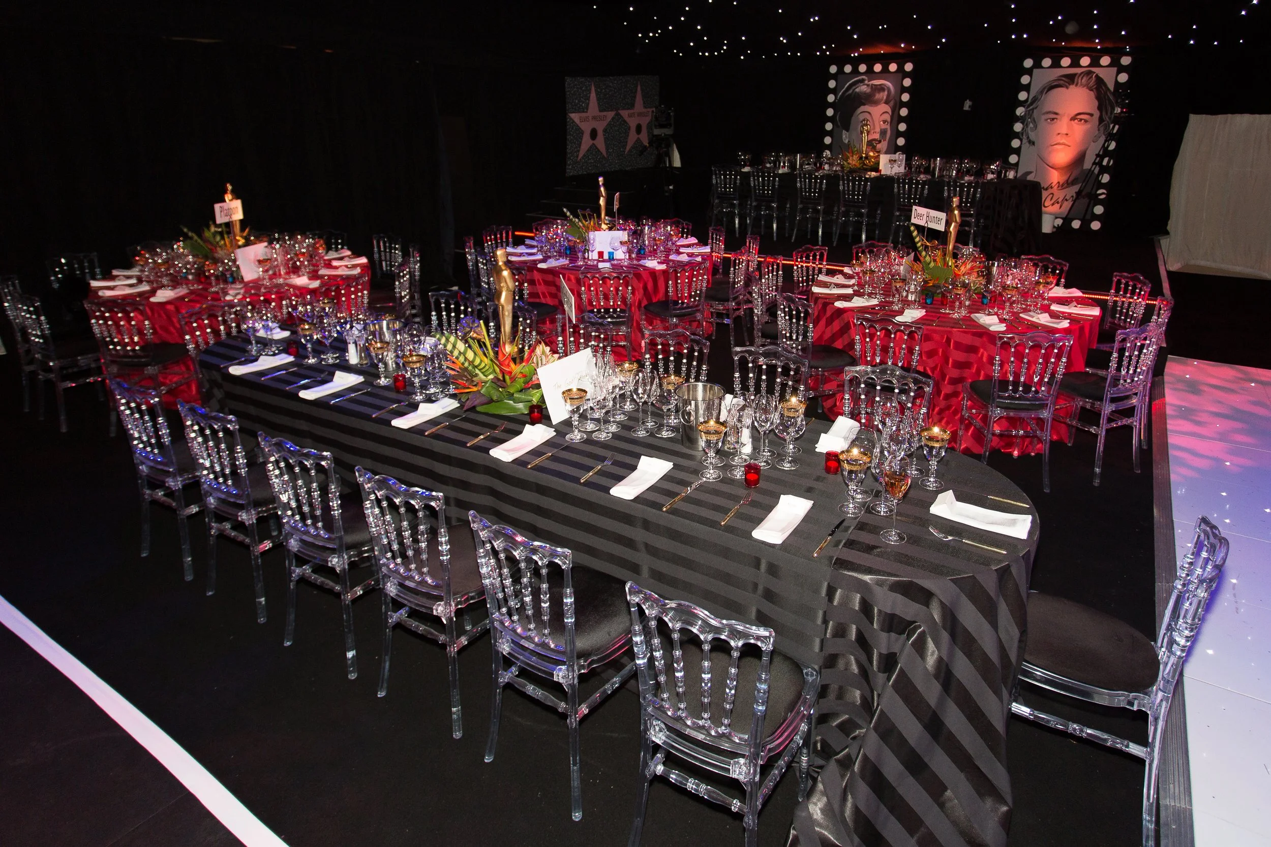Elegantly decorated event space with round tables covered in red tablecloths, surrounded by clear acrylic chairs, set for a formal dinner with glassware and napkins. There are floral centerpieces and place cards on the tables. The background features