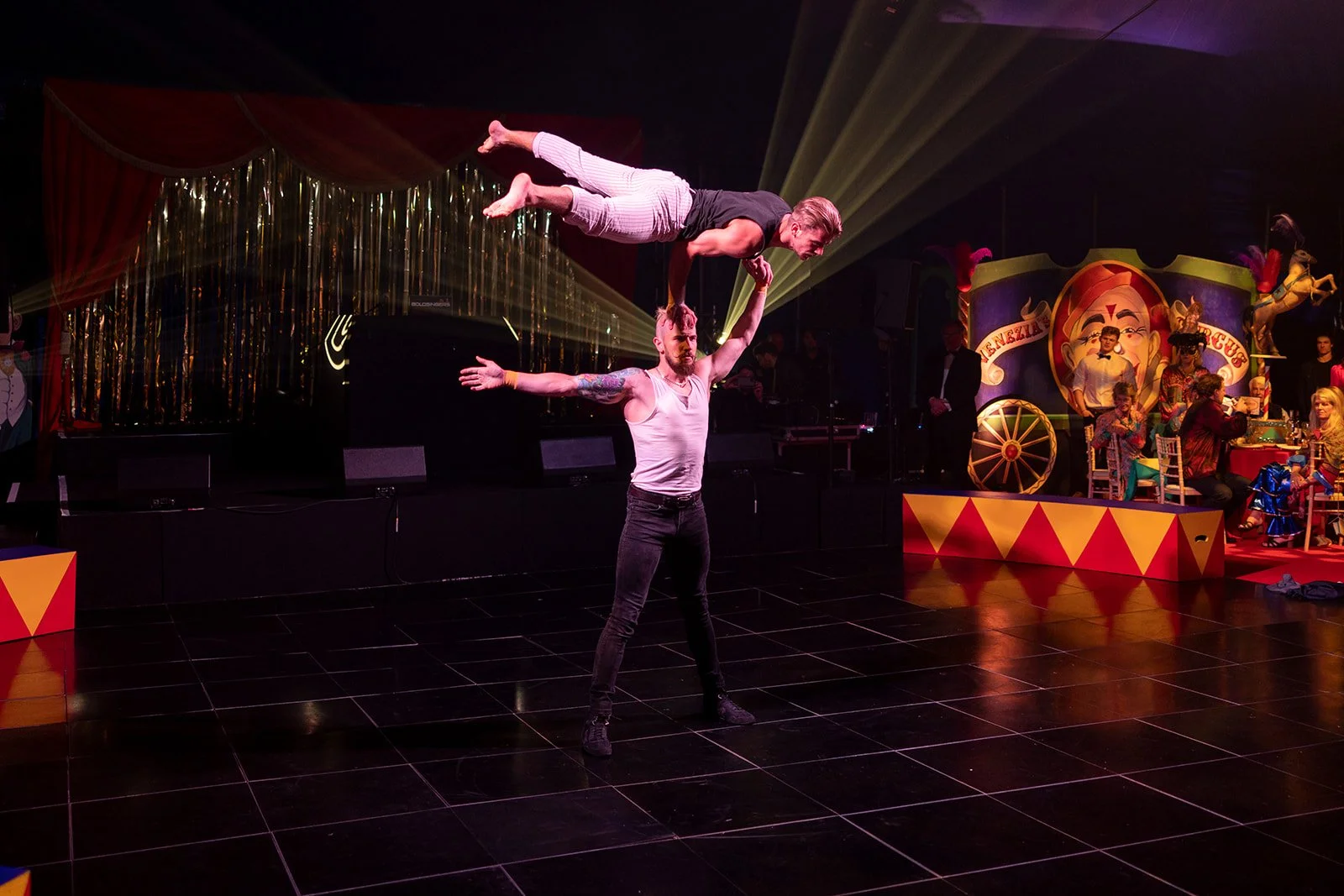 Two performers executing a balancing act on a stage with a circus theme, with an audience and colorful decorations in the background.