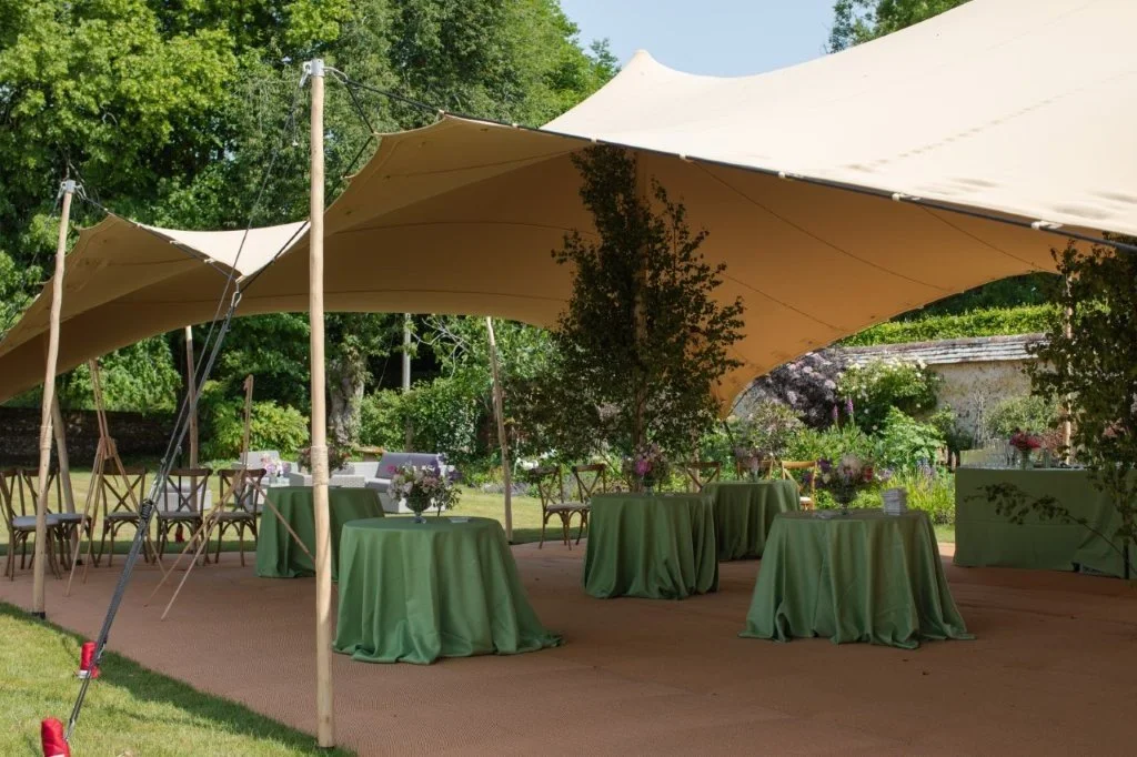 Outdoor event setup with green tablecloths, floral centerpieces, and large beige canopy tents in a garden setting.