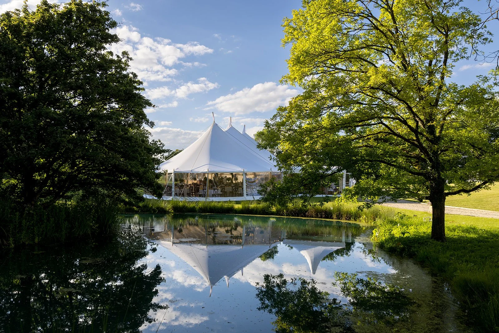 A large white event tent with clear sides is set up in a grassy area near a pond, surrounded by trees, with a clear blue sky overhead.