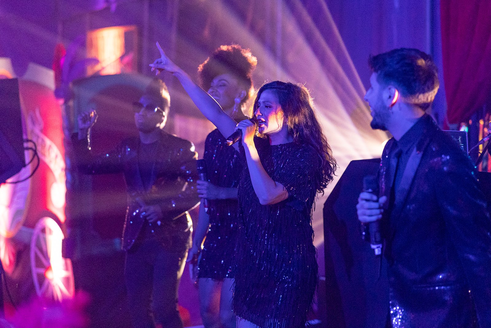 A woman in a black sequin dress is singing into a microphone while pointing upward, surrounded by people in a concert or party setting with colorful lighting.