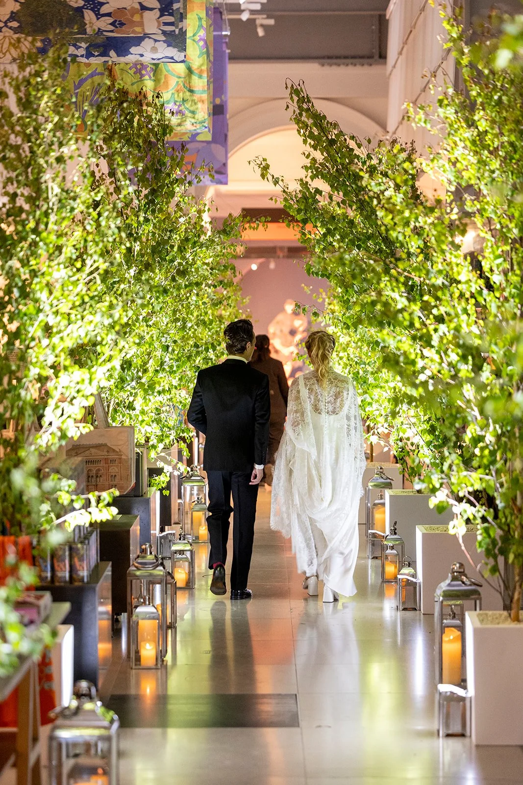 A bride and groom walking together down an aisle decorated with green foliage and lit lanterns inside a venue.