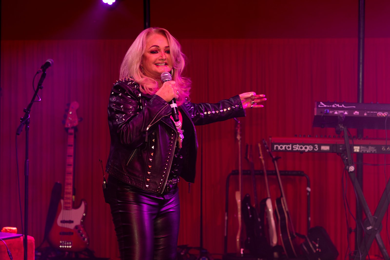 Blonde woman singing on stage in a black leather jacket with studded details, holding a microphone, with musical instruments behind her including guitars and keyboards.