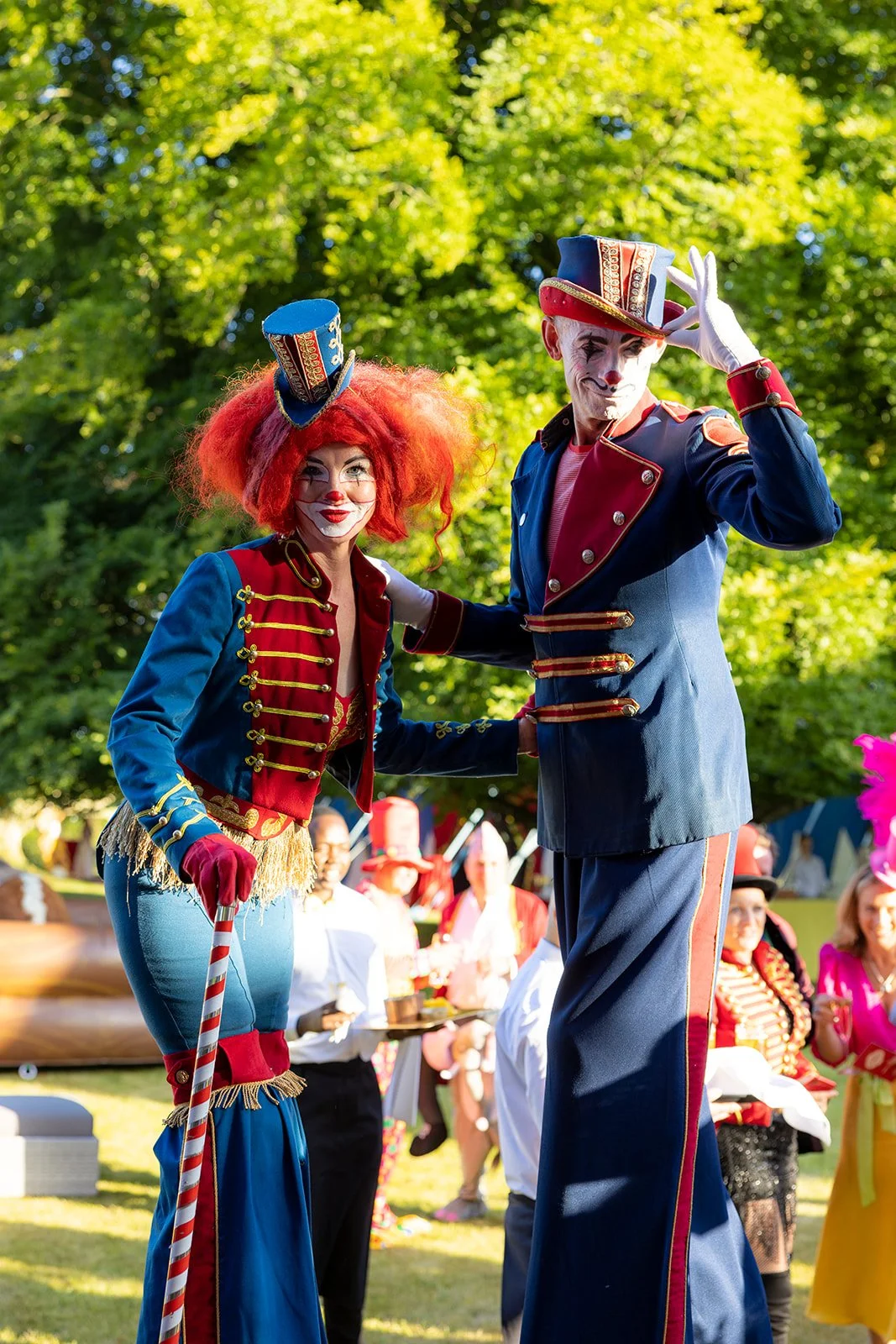 Two performers dressed as circus clowns in colorful costumes and face paint, standing outdoors during a parade or festival with green trees and spectators in the background.