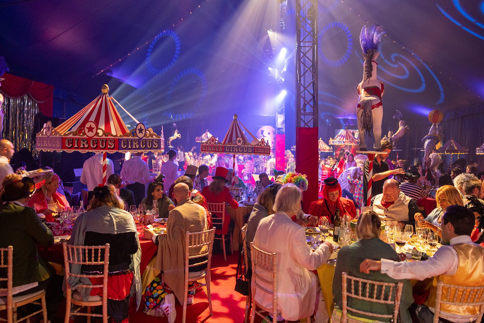A lively circus-themed banquet with guests in colorful costumes seated at tables, surrounded by circus tents, acrobats, and performers on stage, with vibrant lighting and festive decorations.