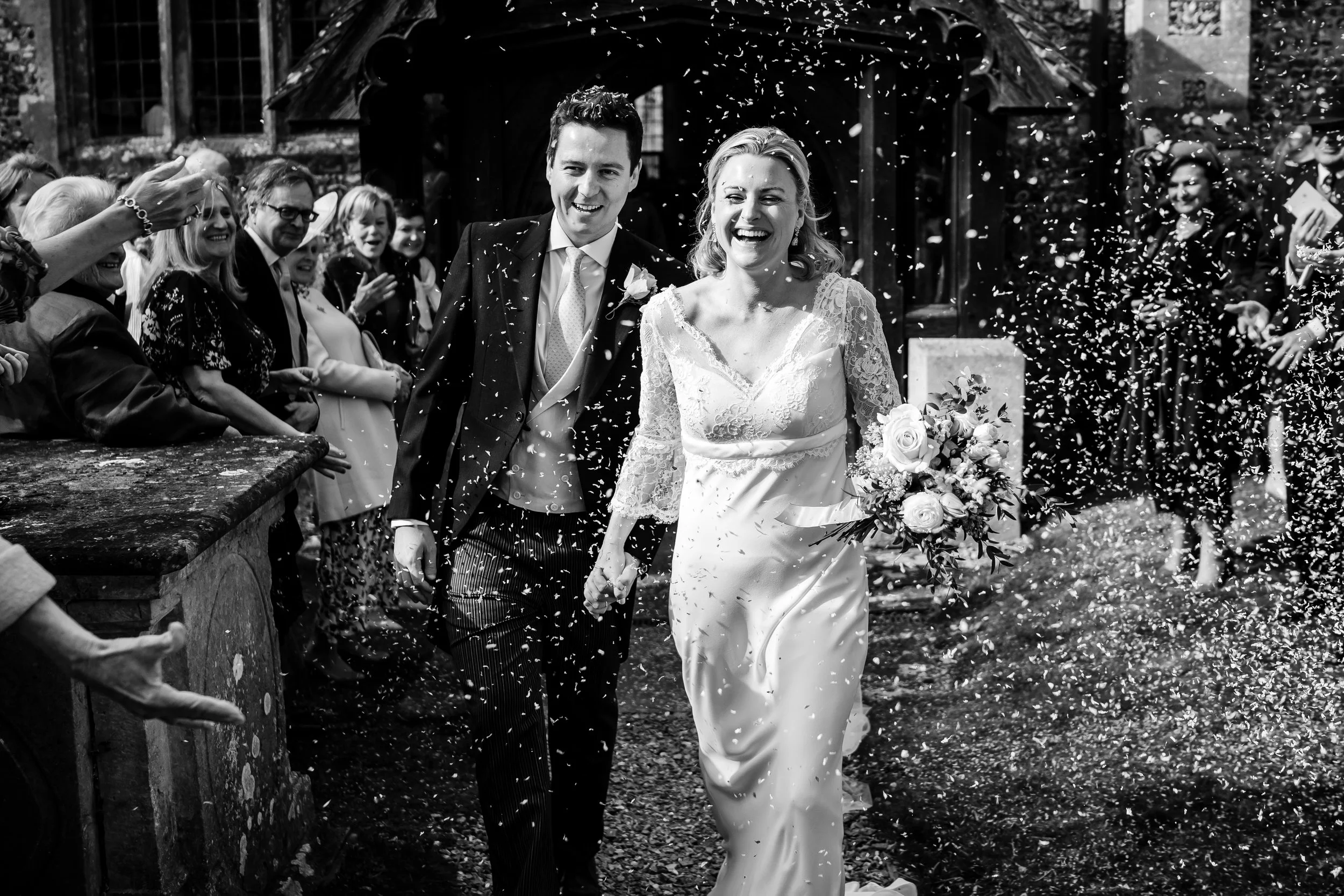 Black and white photo of a bride and groom walking outdoors, surrounded by guests, with confetti falling around them after their wedding ceremony.