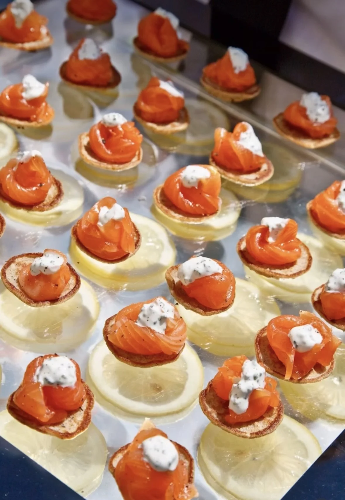 Close-up of canapé bites with smoked salmon on cream cheese on round crackers topped with dollops of white sauce and black pepper, arranged on a baking sheet.