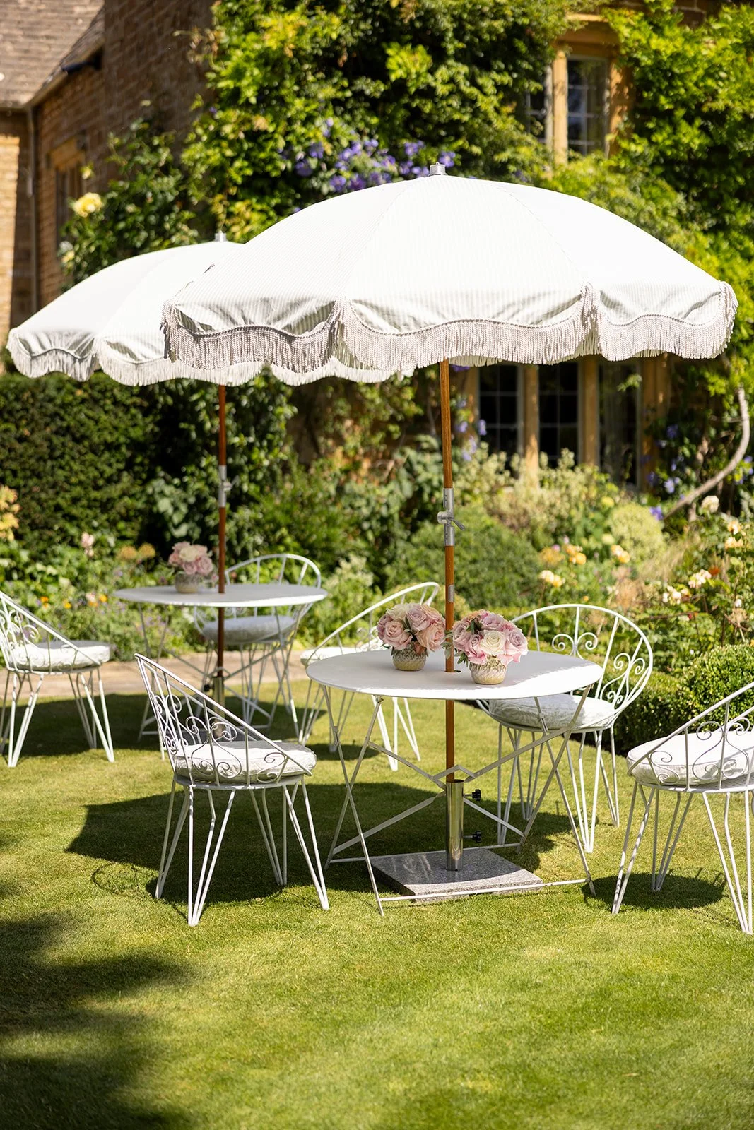 White outdoor patio table with a large white umbrella, surrounded by white metal chairs with cushions, on a green lawn with bushes and a house in the background, decorated with pink flowers on the table.