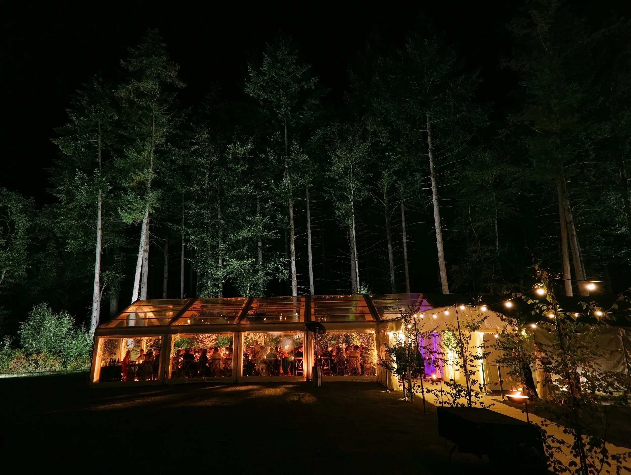 Night scene of an outdoor gathering under a glass structure with warm lighting. In the background, tall trees are visible against the dark sky. Several people are seated and mingling inside the illuminated structure.