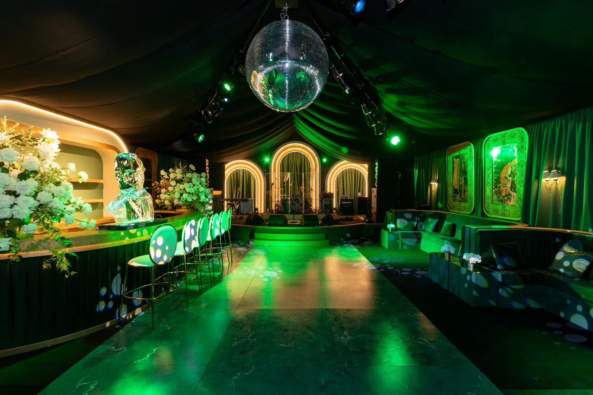 Empty event space with a disco ball hanging from the ceiling, green lighting, floral decorations, a bar area with illuminated stools, musical instruments on stage, and decorated walls with large mirrors.