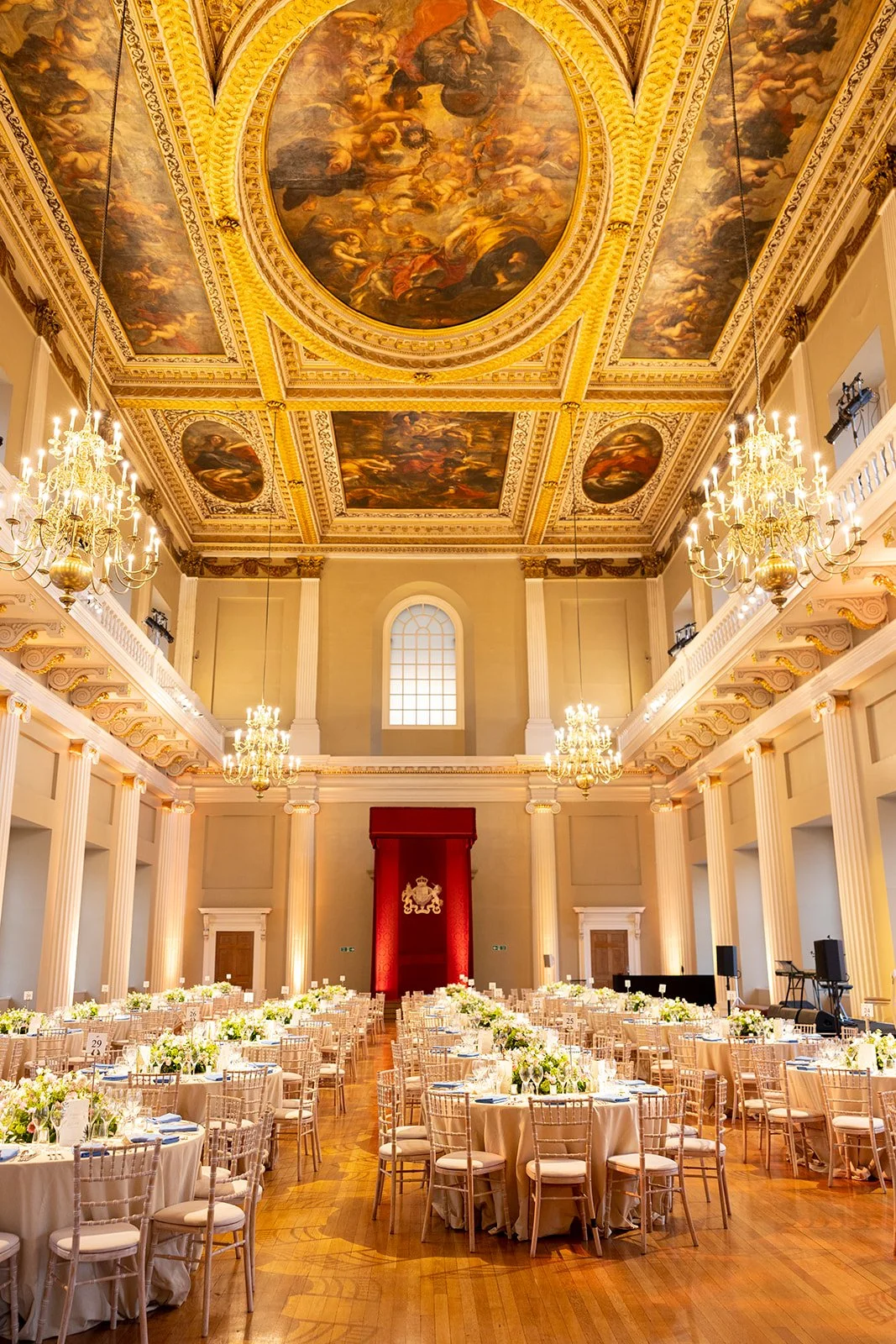Elegant banquet hall with high ceilings, ornate chandeliers, and a decorated ceiling with paintings. Long tables with white tablecloths, flower centerpieces, and set dining ware. A large red drape with a crest at the front of the room.