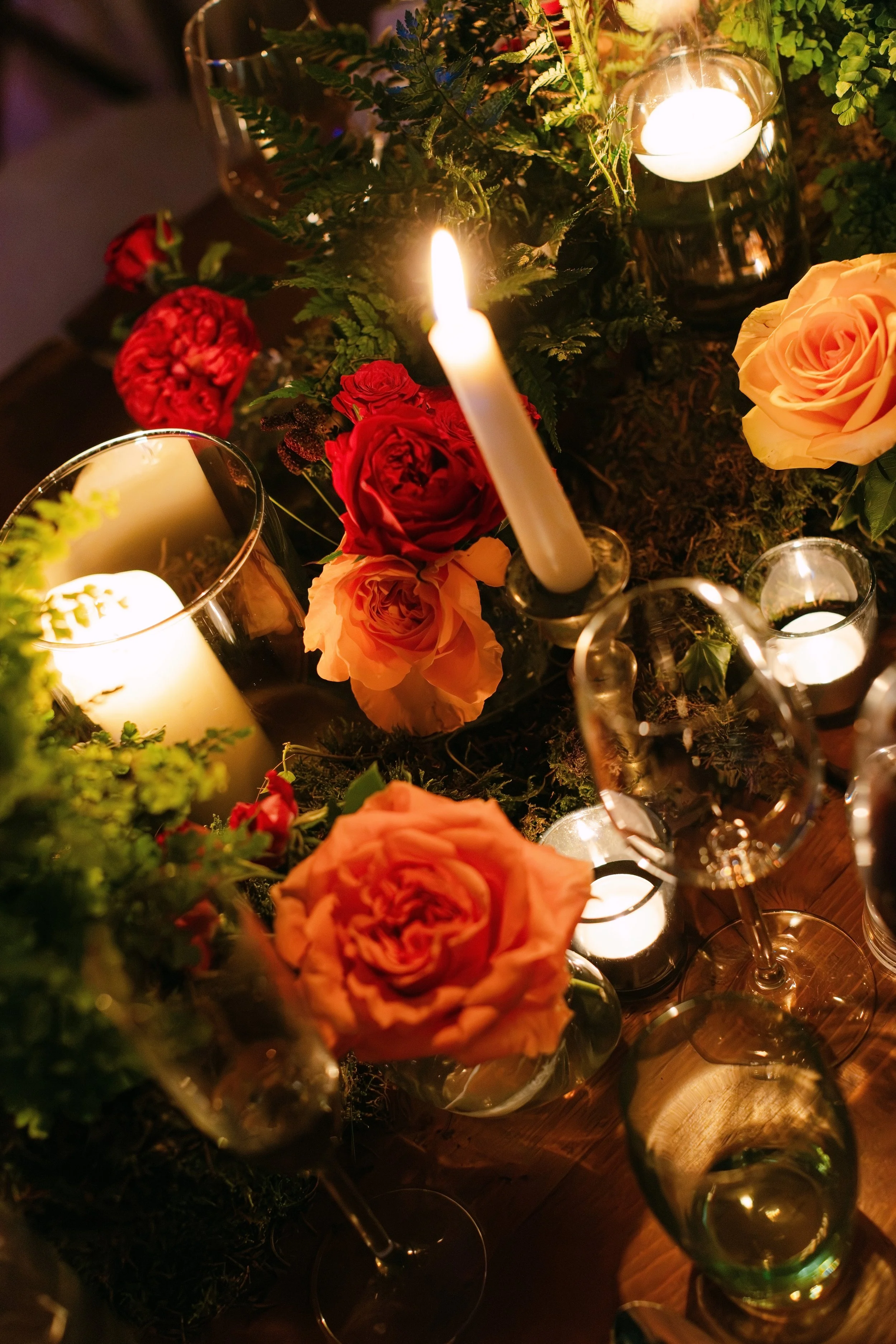 A close-up view of a floral arrangement with roses and greenery, candlelight flickering, and glass candle holders and wine glasses.