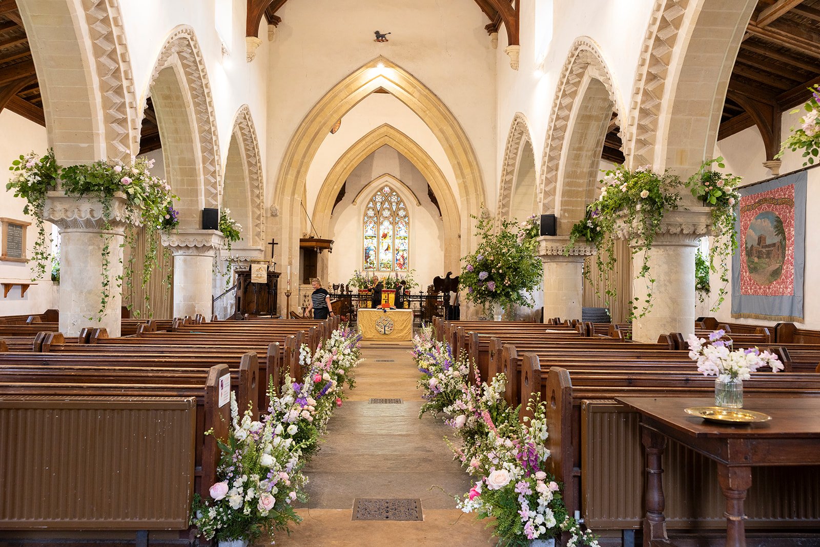 Interior of a wedding chapel decorated with flowers, with wooden pews on either side of a central aisle, a small altar at the front, and stained glass windows.