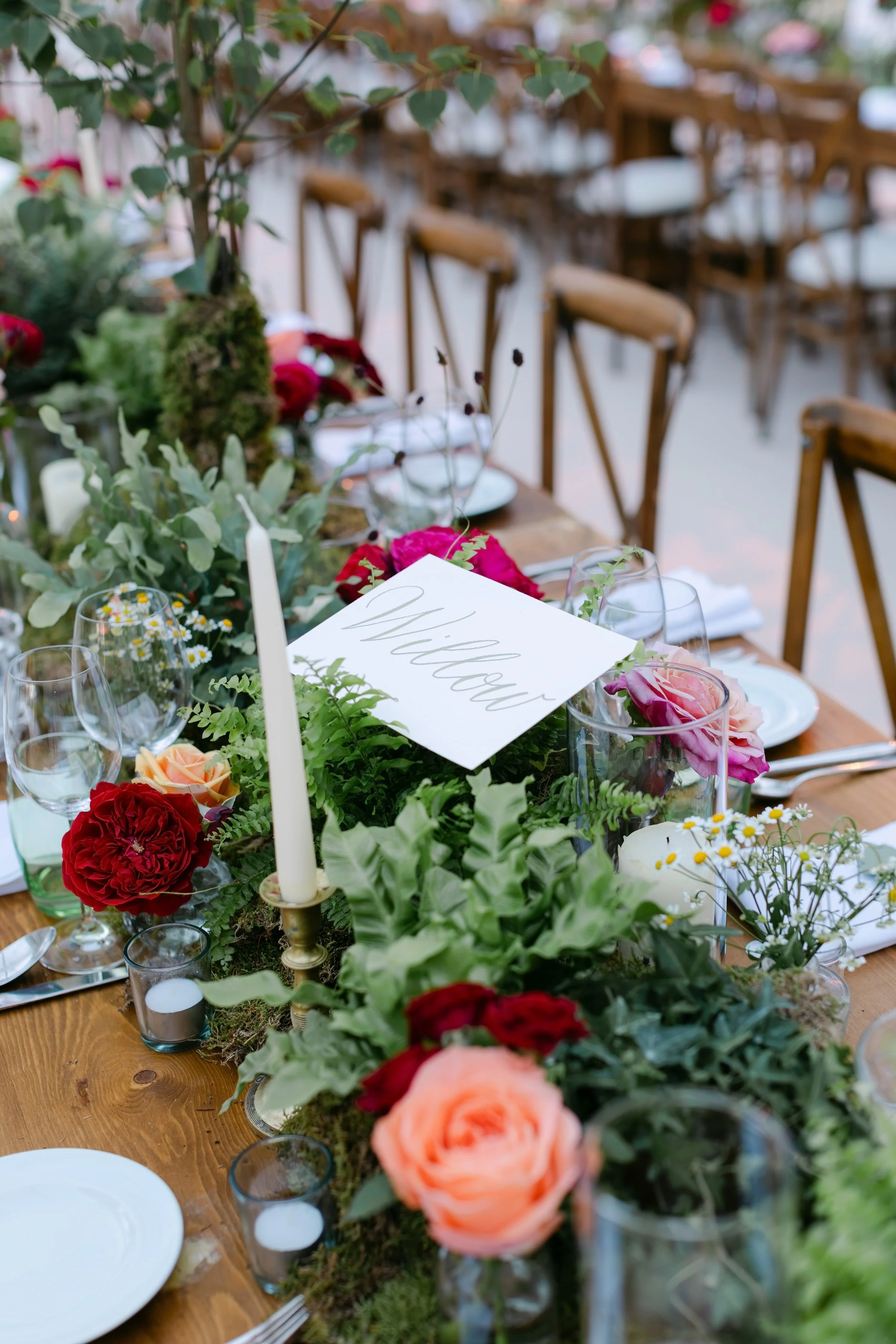 A decorated dining table with a floral centerpiece, candles, glassware, and a 'Thank You' card. Wooden chairs are arranged around the table in an indoor or outdoor setting.