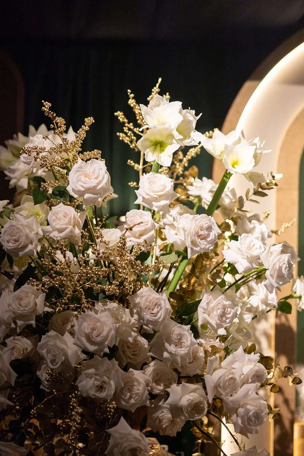 Close-up of a floral arrangement with white roses, white lilies, gold decorative accents, and greenery in front of a dark background with curved architectural elements.