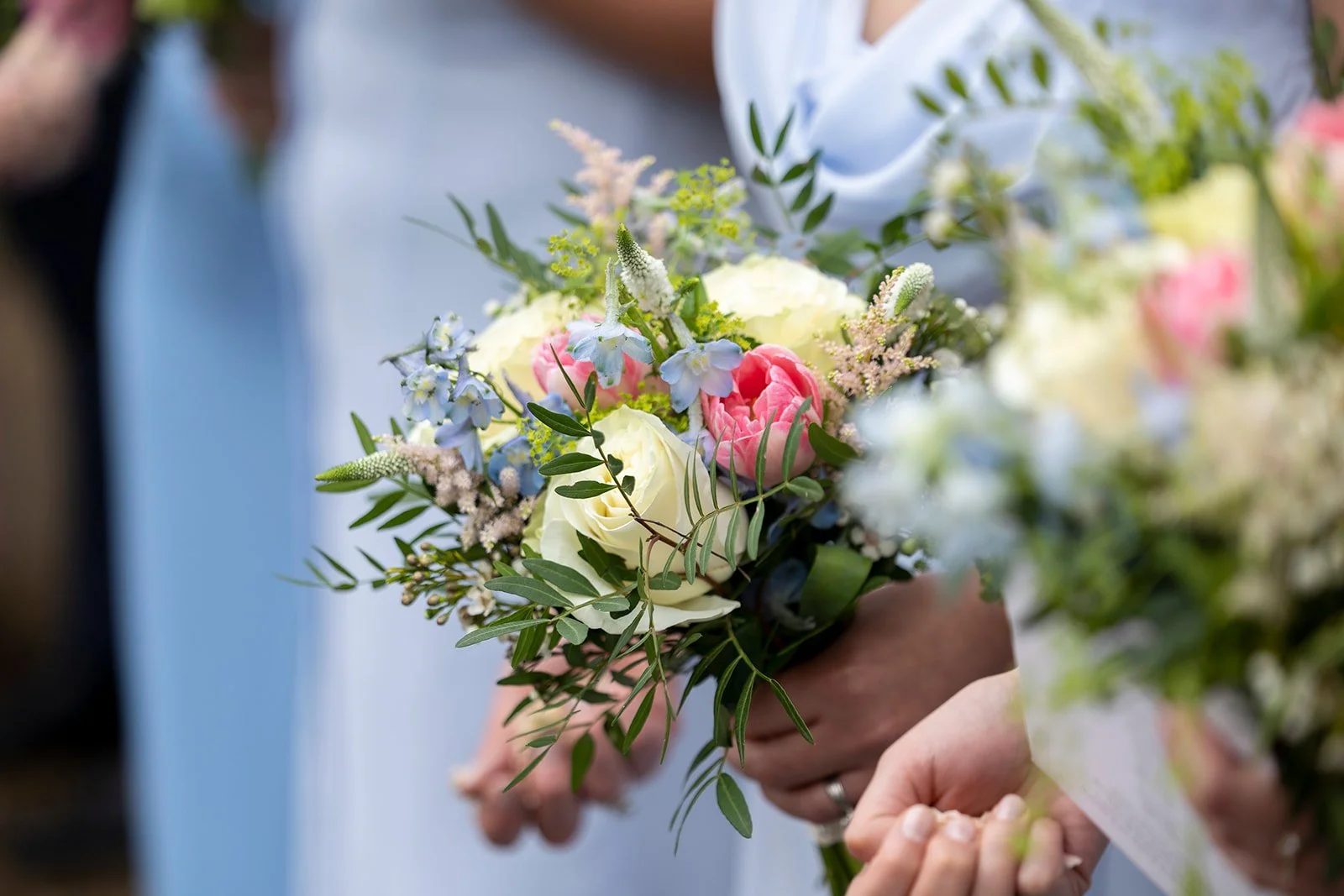 Close-up of a person holding a wedding bouquet with white, pink, and blue flowers and green leaves.