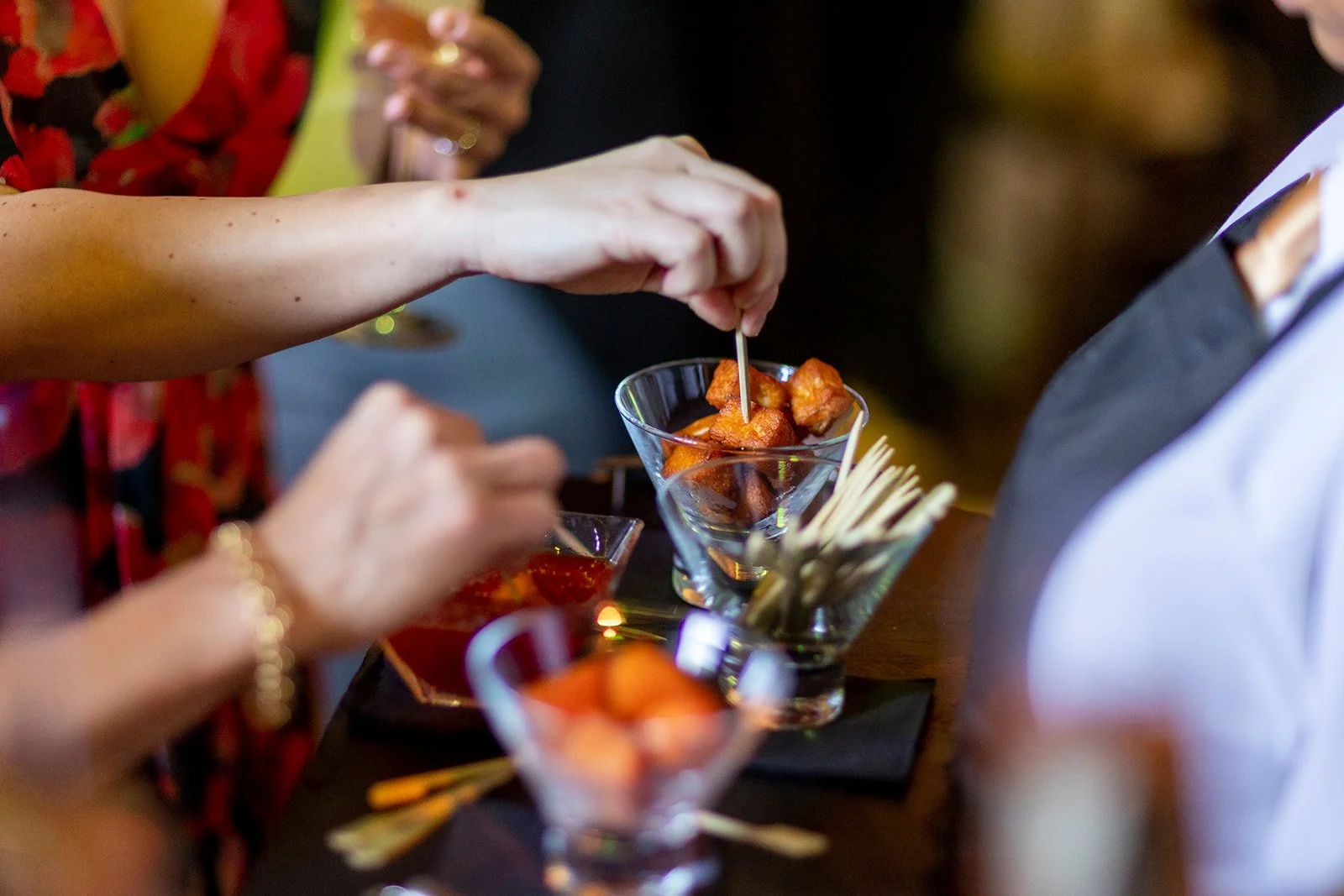 People sharing appetizers at a social gathering, with focus on a hand skewering food in a martini glass filled with fried appetizer cubes, and others holding drinks in the background.