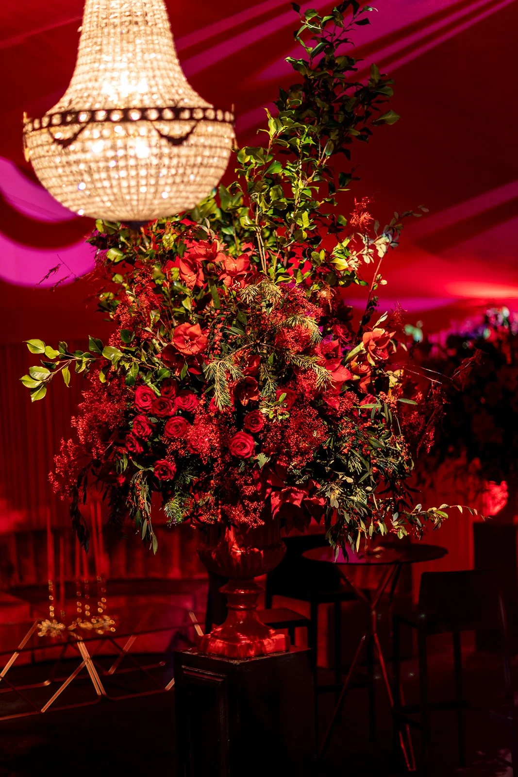 A floral arrangement with red roses, green leaves, and red flowers in a large vase on a black pedestal, illuminated with red lighting, with a crystal chandelier hanging above.