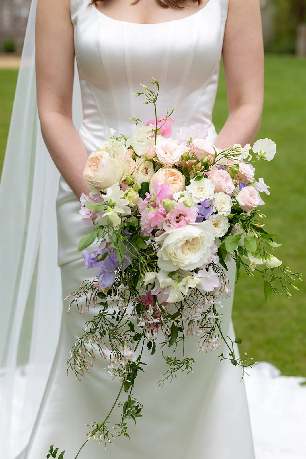 A bride in a white satin wedding dress holding a cascading bouquet of pink, white, and purple flowers outdoors.