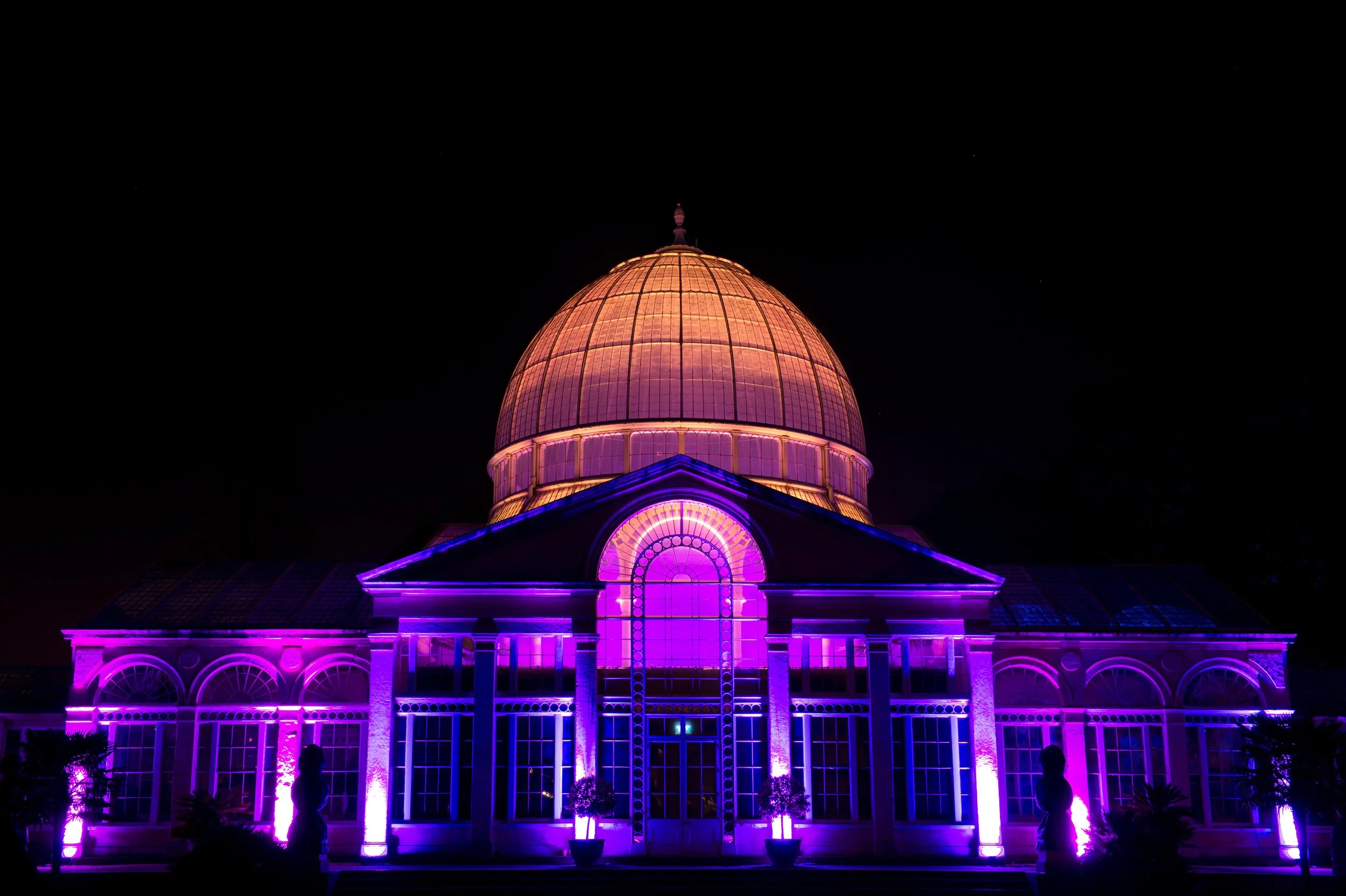 A building with a large domed roof illuminated in pink, purple, and orange lights at night. The architecture features arched windows and decorative elements, with silhouettes of people in front.