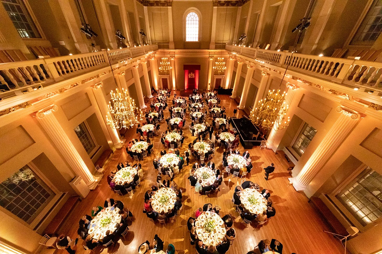 Elegant banquet hall with high ceilings, chandeliers, and tall columns, filled with round tables decorated with floral centerpieces, hosting formally dressed guests at a social event.