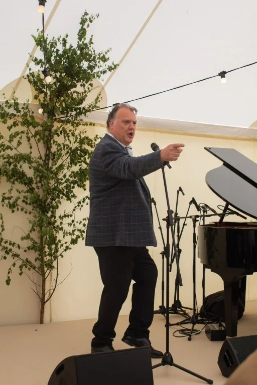 A man in a gray blazer and black pants is standing on a stage, pointing and speaking into a microphone. There is a grand piano to his right and a potted tree behind him. The stage is inside a tent with string lights overhead.