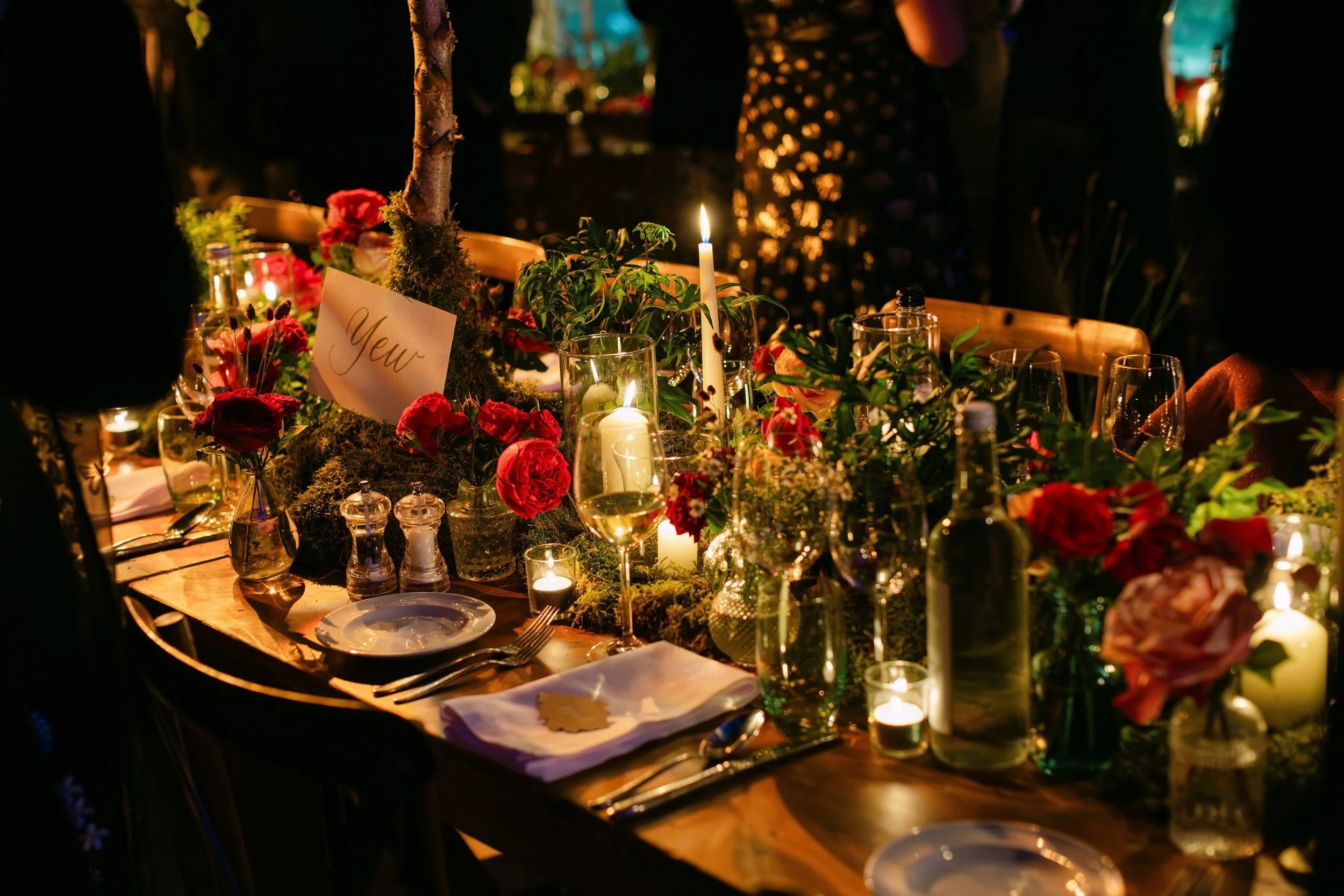 A dimly lit banquet table decorated with flowers, candles, and greenery, set for a special occasion or dinner party.